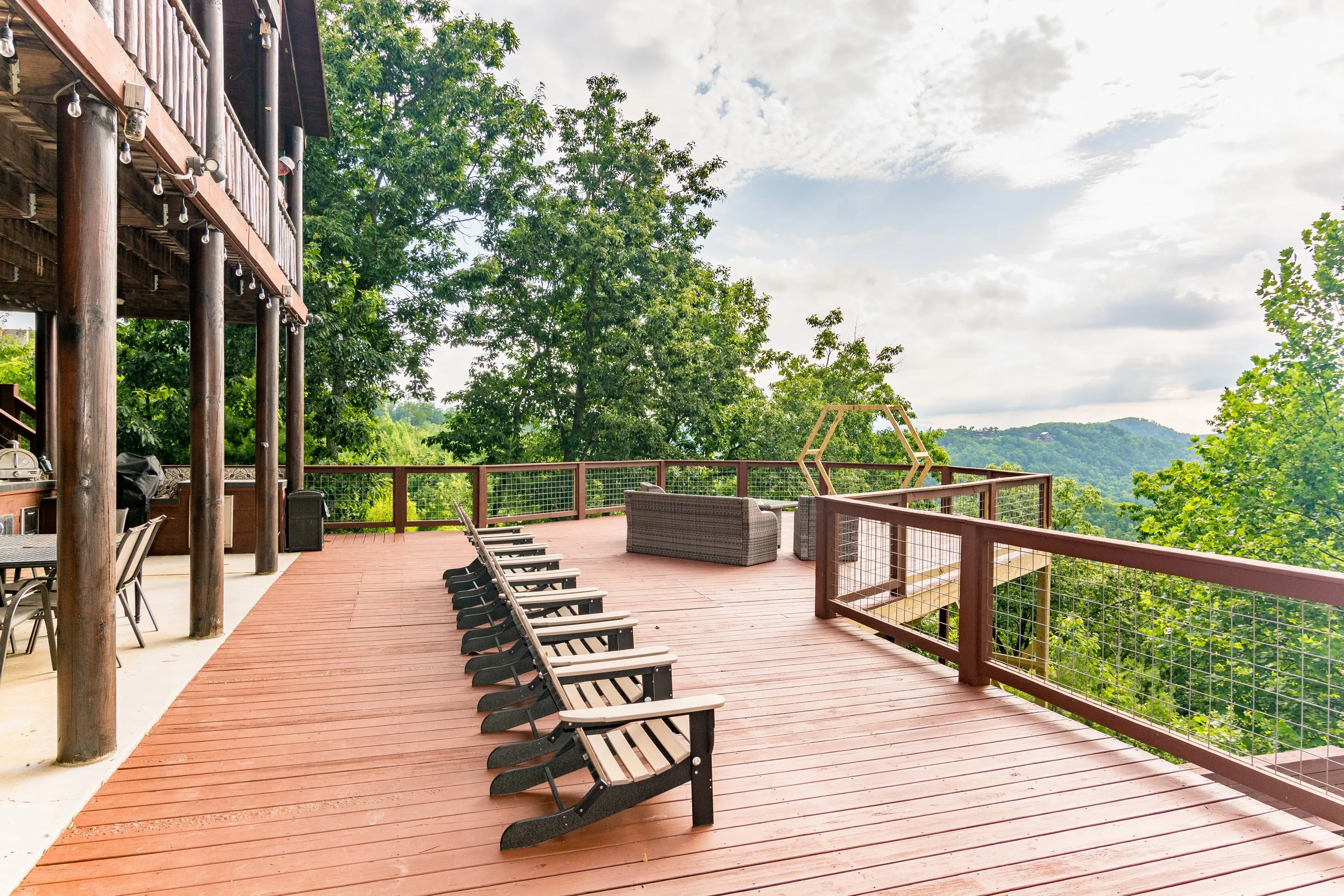 Large outdoor terrace with wooden floorboards, lounge chairs, and a seating area, overlooking green trees and distant hills under a partly cloudy sky.