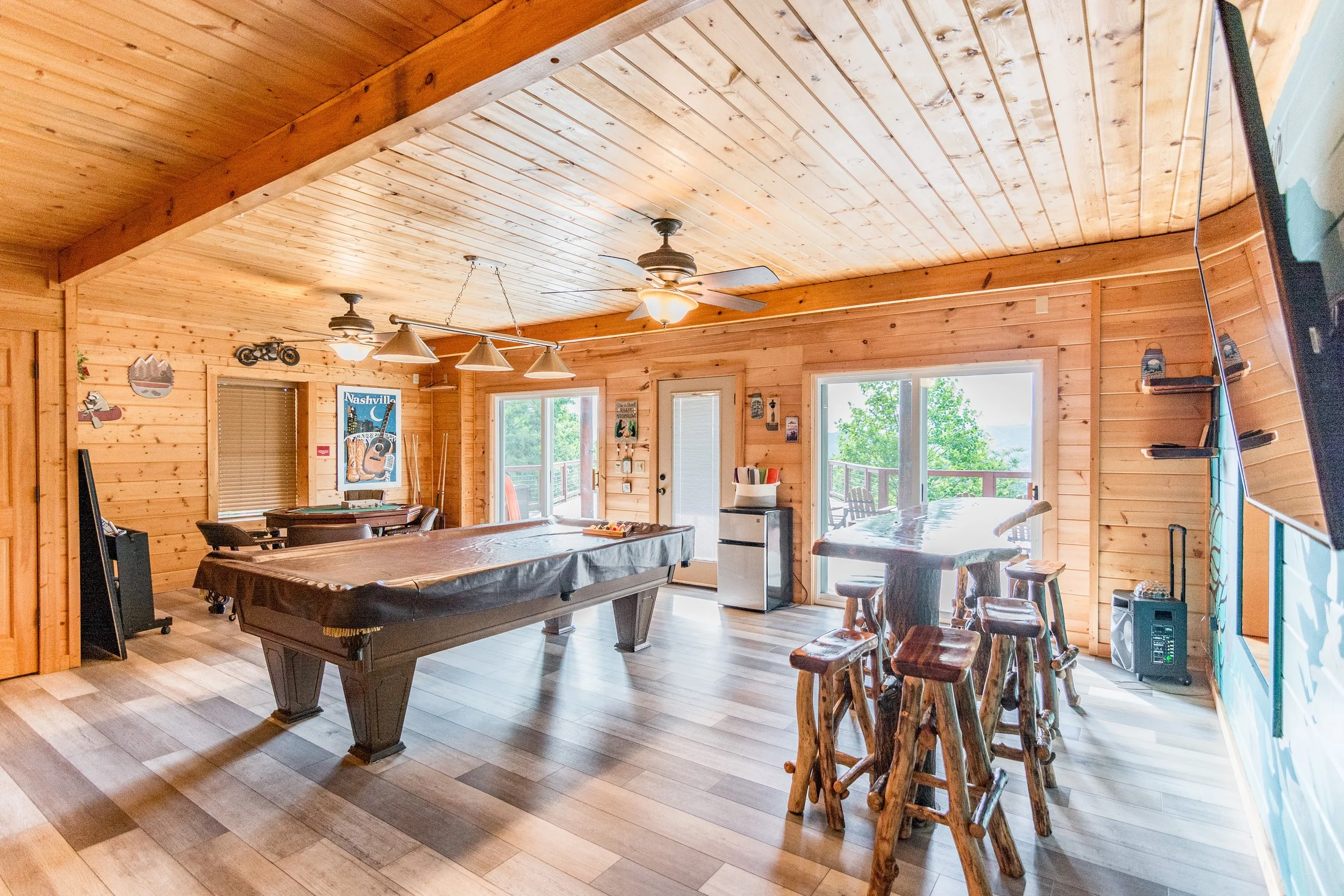 Interior view of a wooden cabin game room with a pool table, bar stool seating, a patio door, and windows showing trees outside.