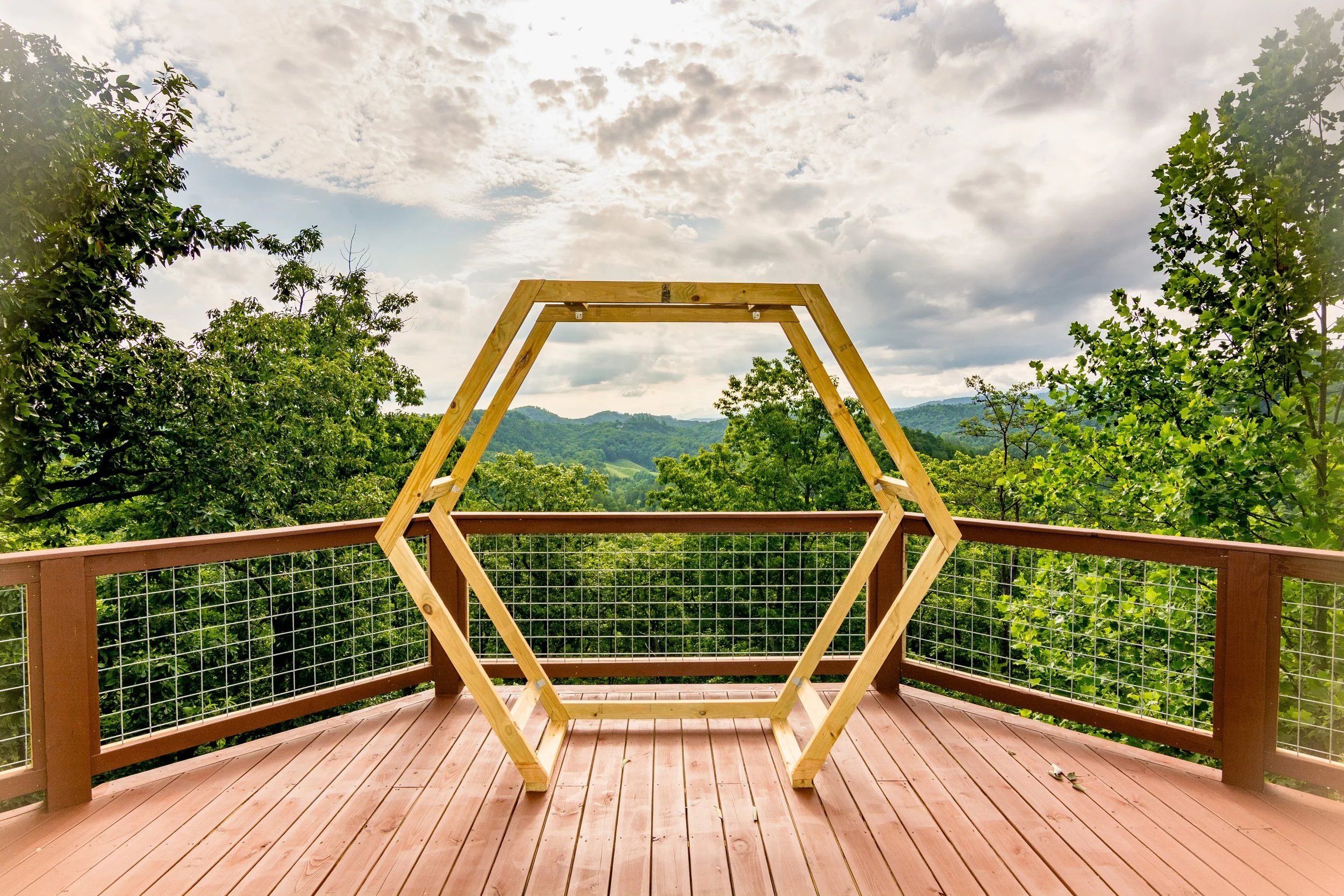 A wooden hexagonal frame on a wooden deck with a scenic view of green trees and mountains under a cloudy sky.