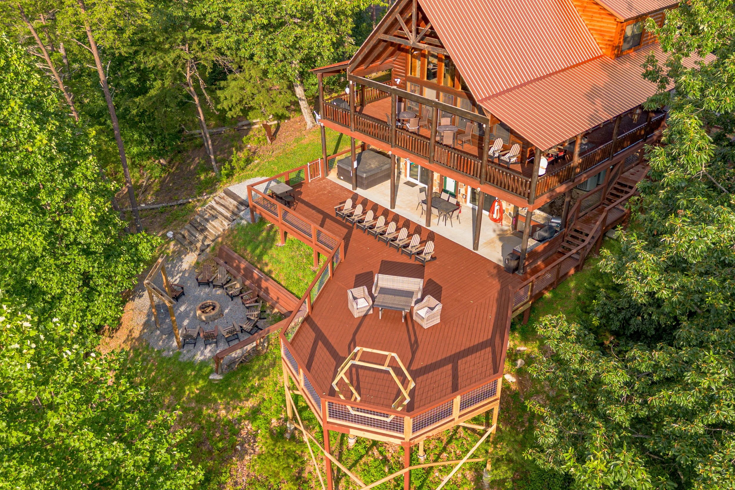 Aerial view of a multi-level wooden house with decks, surrounded by green trees, with outdoor furniture and a fire pit area.