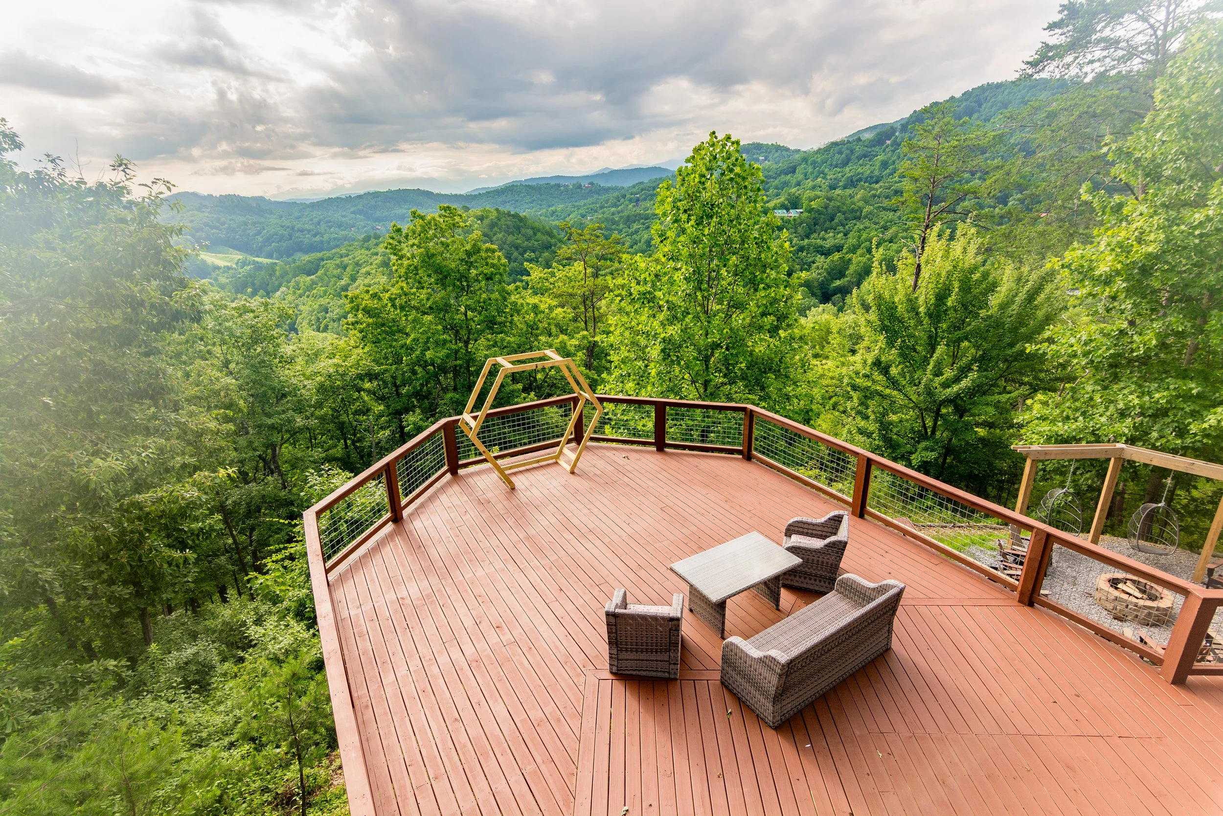 Large wooden deck with outdoor furniture, overlooking a lush green forested mountain landscape under cloudy skies.