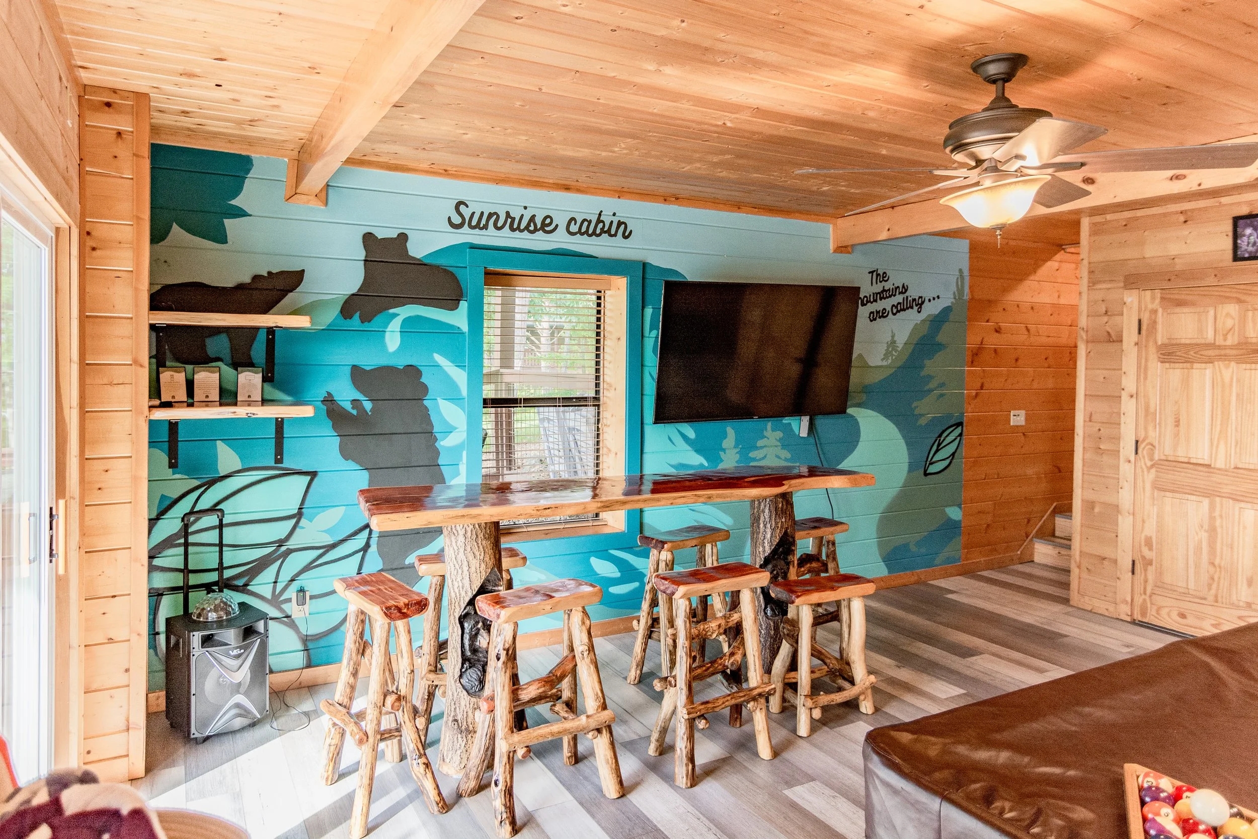 Interior view of a wooden cabin with a colorful wall mural featuring bears and forest scenery, a mounted flat-screen TV, a bar-height table with rustic wooden stools, ceiling fan, and stairs on the right.