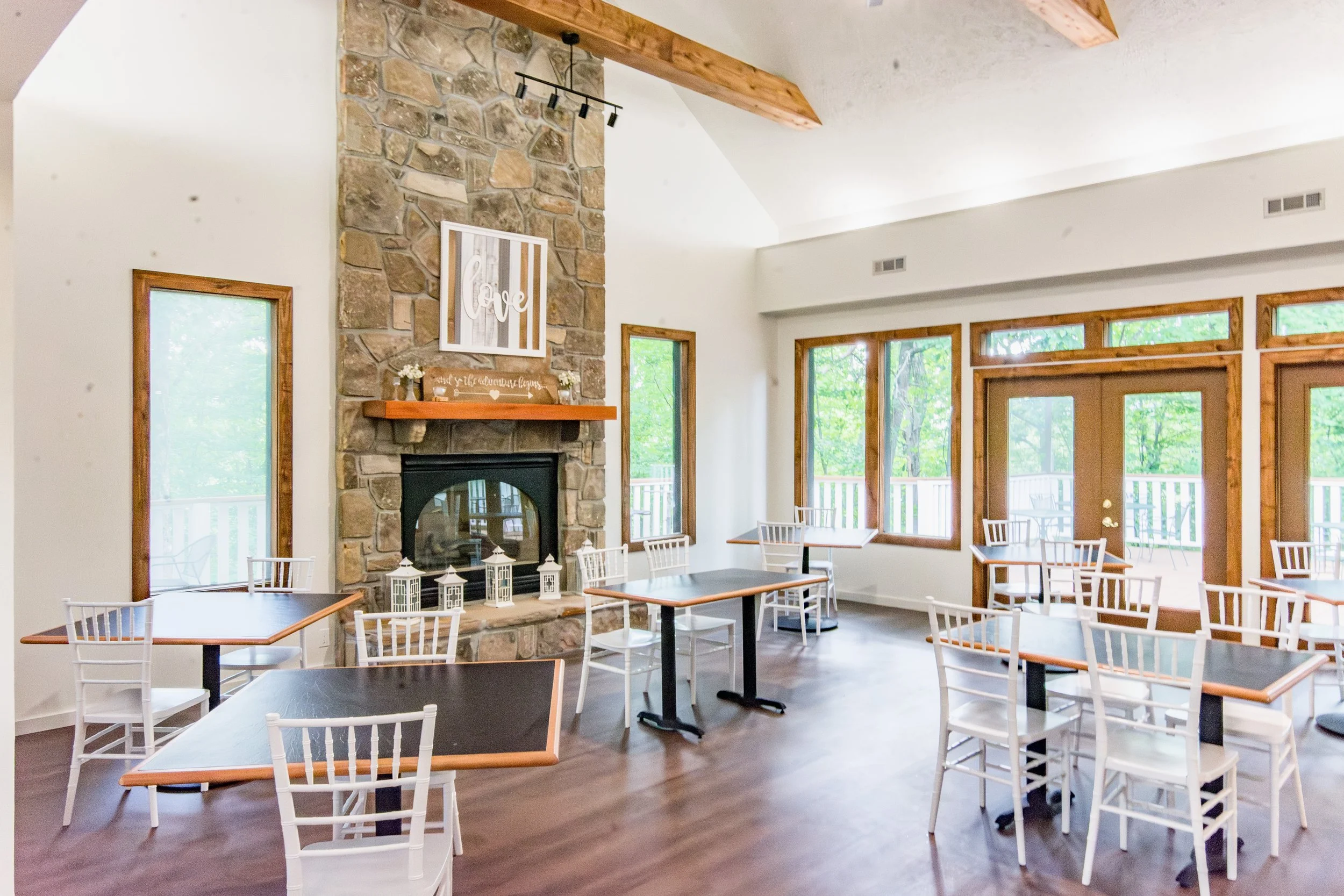 A bright dining area with multiple tables and white chairs, a stone fireplace with a wooden mantle, and large windows and glass doors showing green trees outside. There is a decorative sign on the mantle and a framed art piece with the word 'love' ha