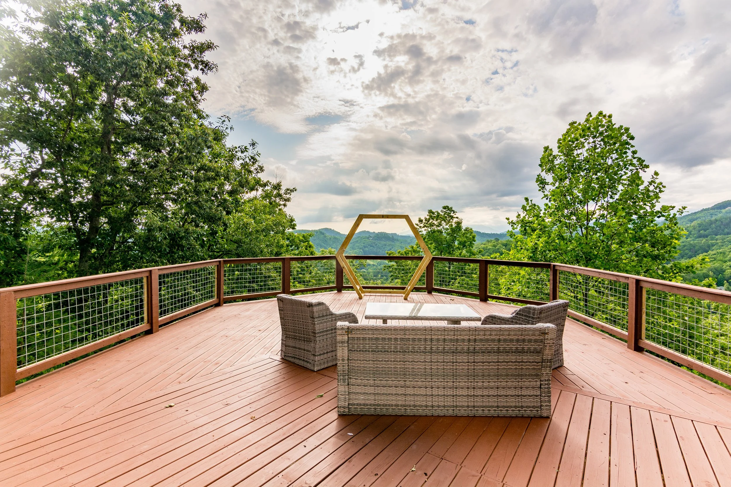 A spacious wooden deck with outdoor furniture overlooking lush green mountains and trees under a cloudy sky.