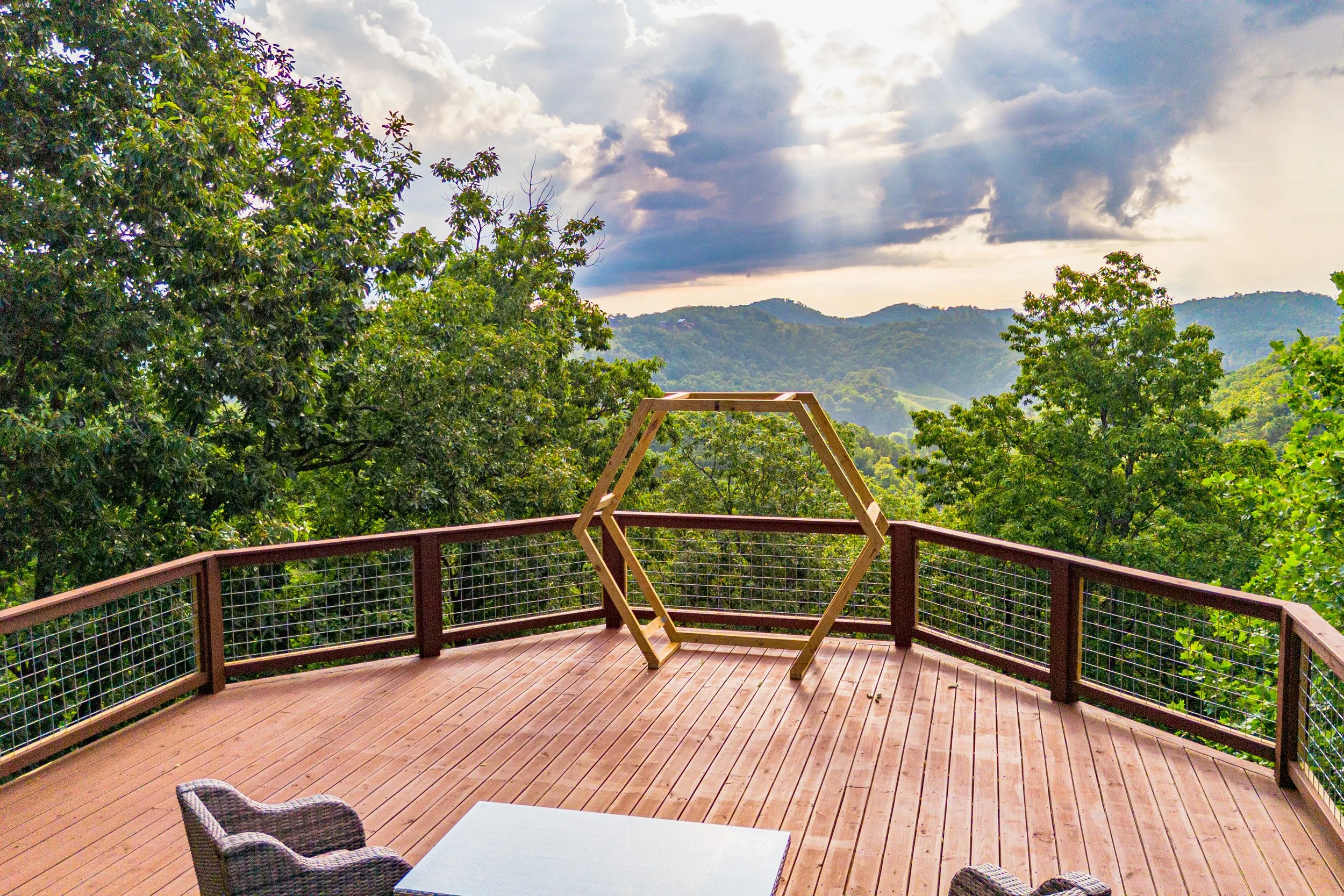 An outdoor wooden deck with a mountain view, surrounded by green trees, cloudy sky with rays of sunlight, a wicker chair, a table, and a hexagonal wooden frame.