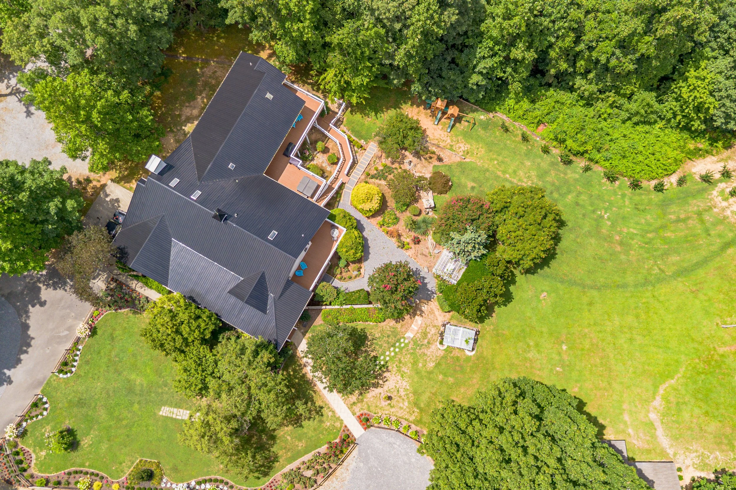 An aerial view of a large house with a dark roof, surrounded by green trees and a spacious yard with sidewalks, garden beds, and outdoor seating areas.