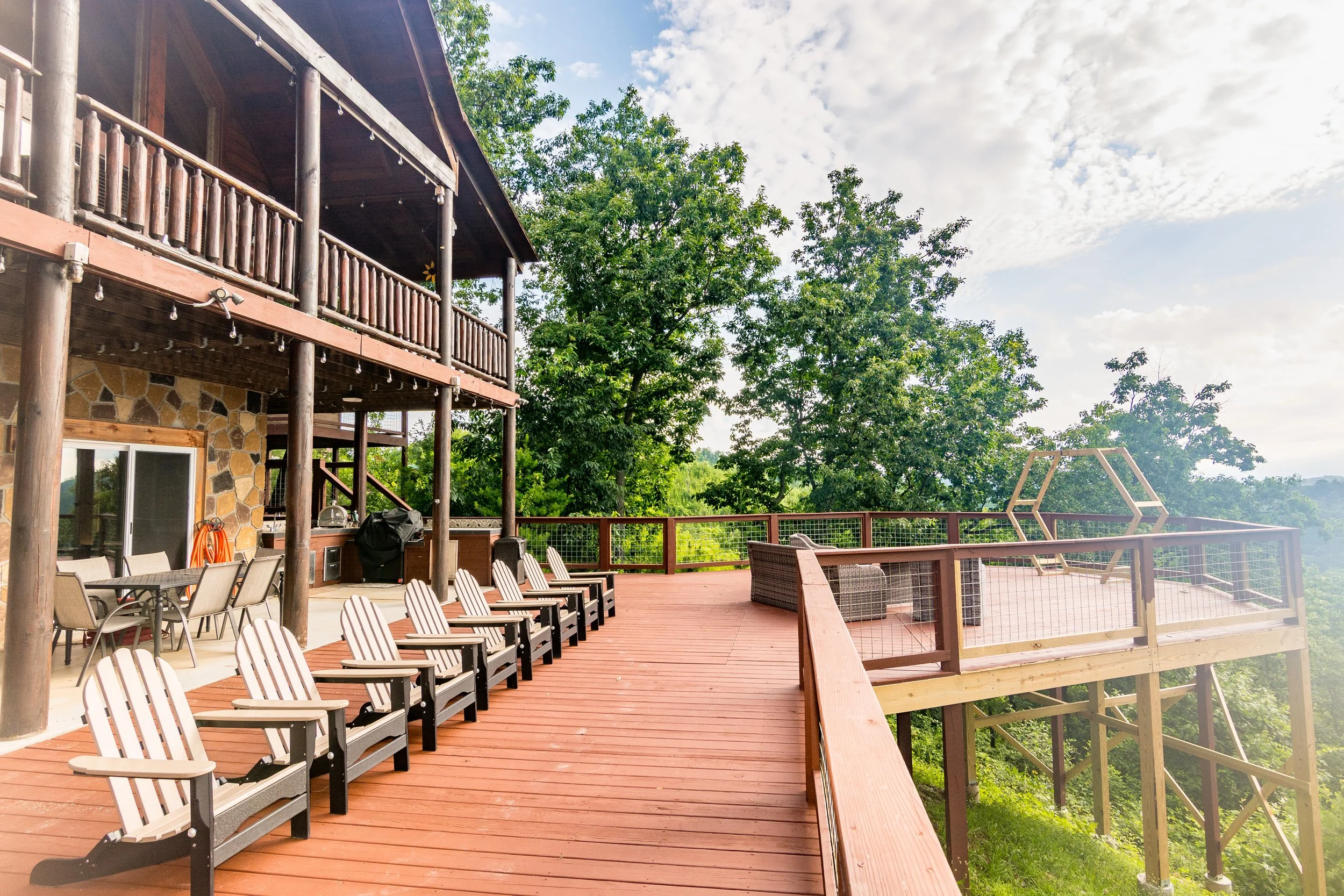 Wooden deck with outdoor furniture, Adirondack chairs, and a tree house structure, surrounded by green trees under a partly cloudy sky.