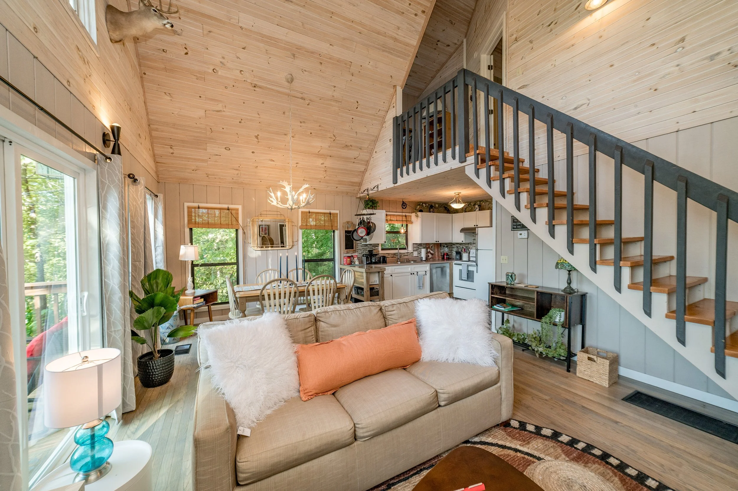 Interior of a cozy living room with wood-paneled walls and ceiling, featuring a beige sofa with orange and white fluffy pillows, a small side table with a turquoise lamp, a potted plant, a dining table with chairs, a kitchen area with white cabinets and appliances, and a staircase with a blue railing leading to an upper level.