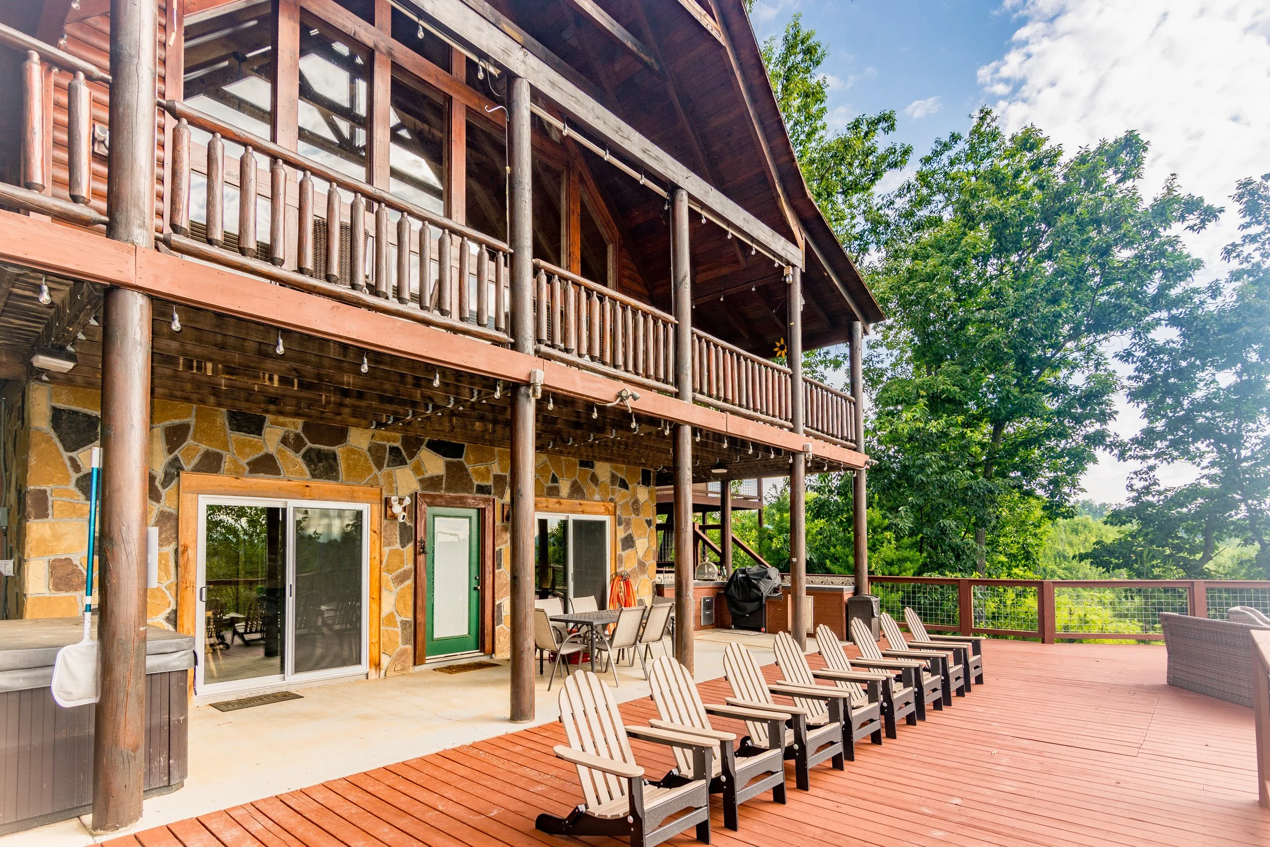 A large wooden house with stone accents and a spacious deck featuring several Adirondack chairs and outdoor furniture, surrounded by greenery and trees.