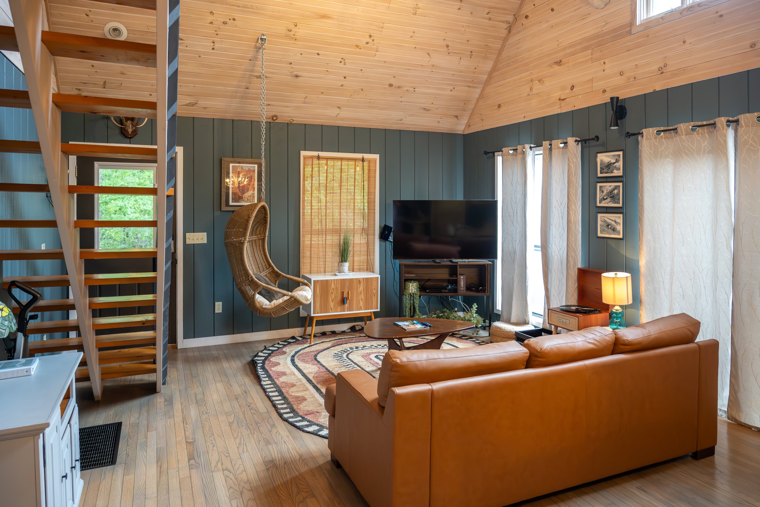 Living room with a tan sofa, a large flat-screen TV, a hanging wicker chair, and wood-paneled ceiling and walls with windows and curtains.