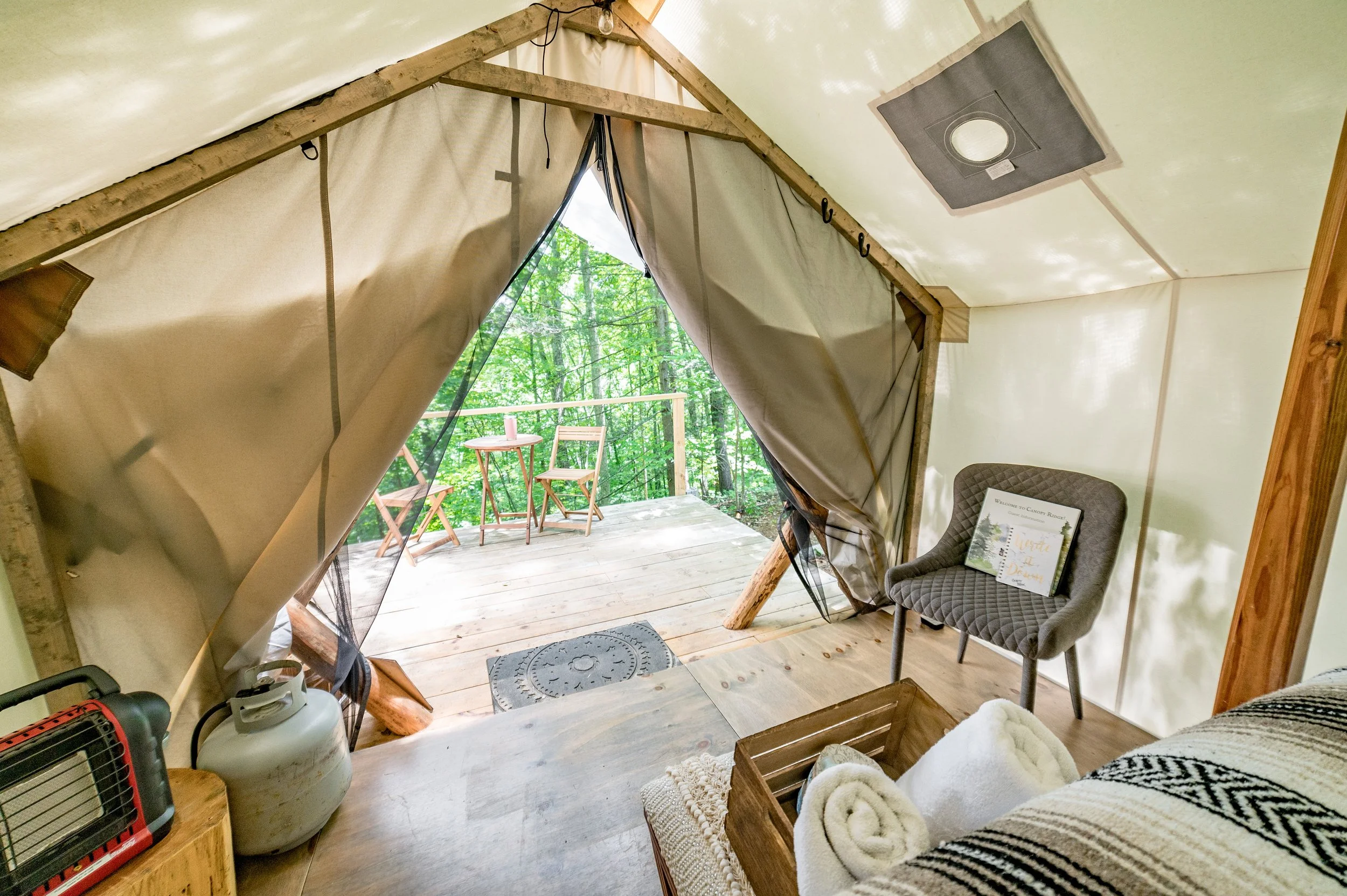 Inside a glamping tent with a small wooden deck outside. The tent has beige fabric walls and a triangular opening with curtains. Inside, there's a comfy gray chair, a bed, and a wooden crate with rolled white towels. Outside, on the deck, are two wooden chairs and a small round table with a pink cup, surrounded by green trees.