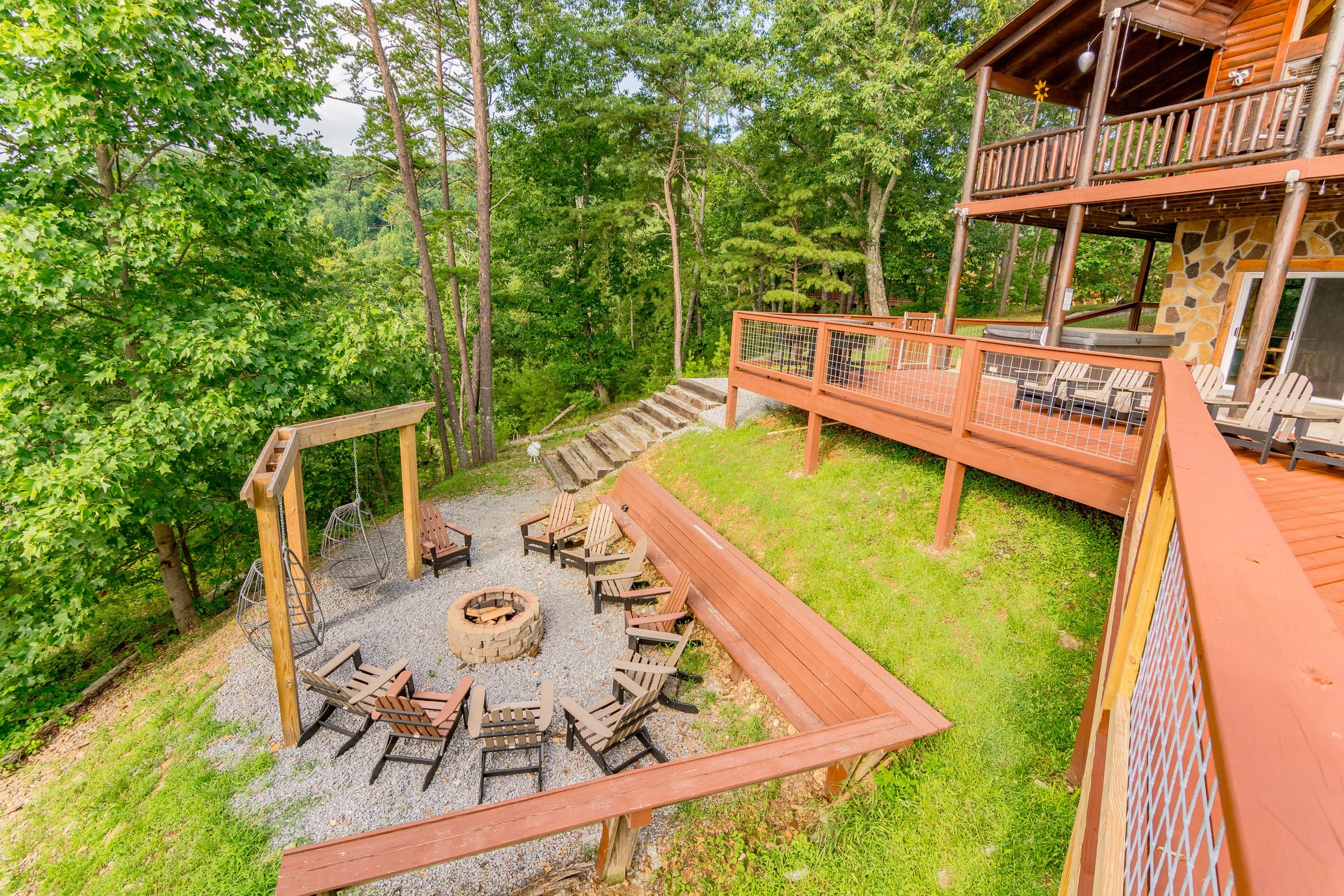 A backyard with a fire pit surrounded by chairs, a wooden swing, and Adirondack chairs on a gravel area, with a tall forest of green trees in the background and a multi-level wooden deck with seating.