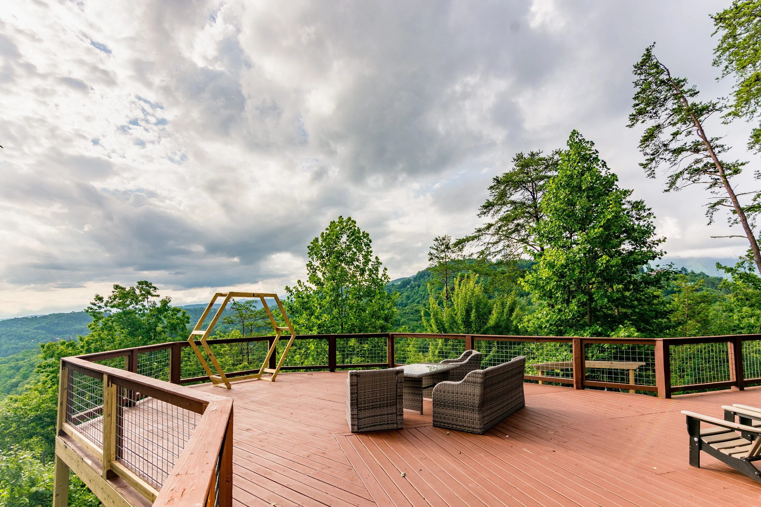 A spacious wooden deck with outdoor furniture, surrounded by green trees and overlooking a hilly landscape under a cloudy sky.