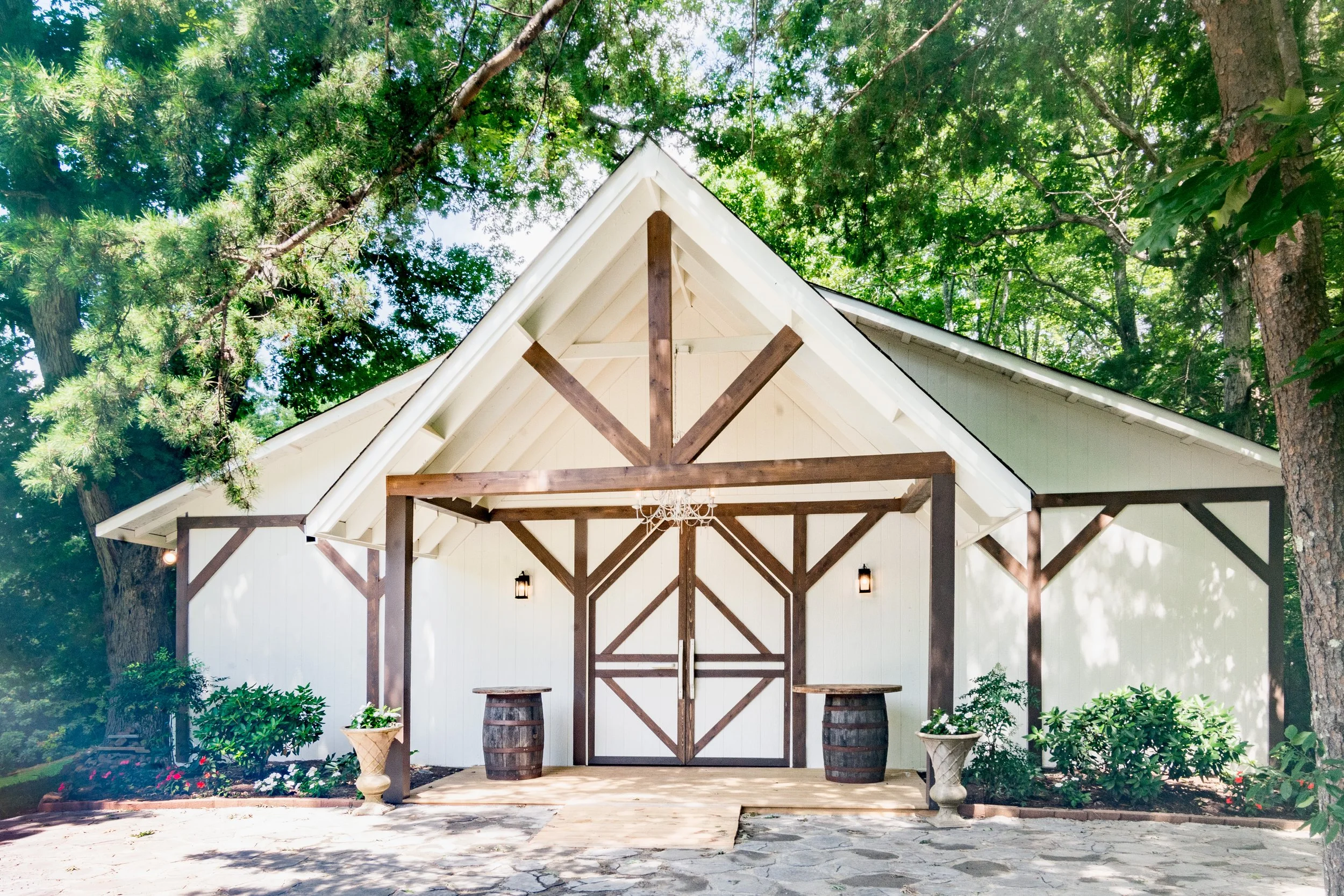 White barn with brown wooden trim and double doors, situated in a lush green wooded area, with potted plants and stone walkway in front.