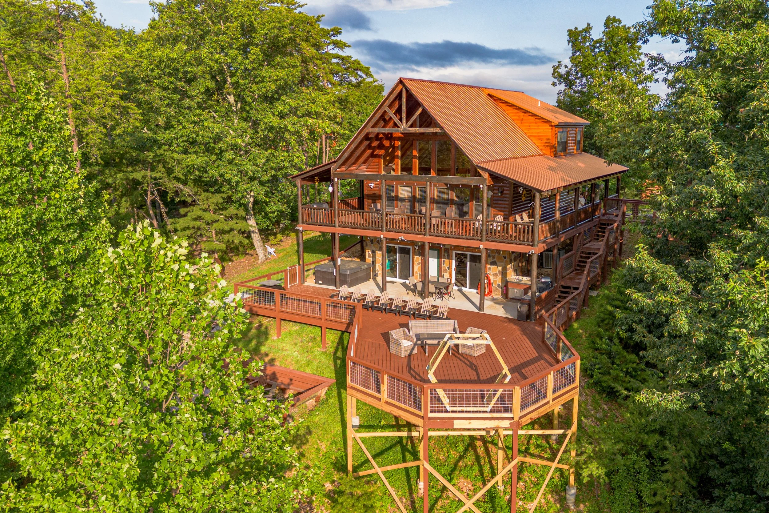 A large multi-story wooden house with a red metal roof, surrounded by green trees, and featuring multiple decks and outdoor furniture.
