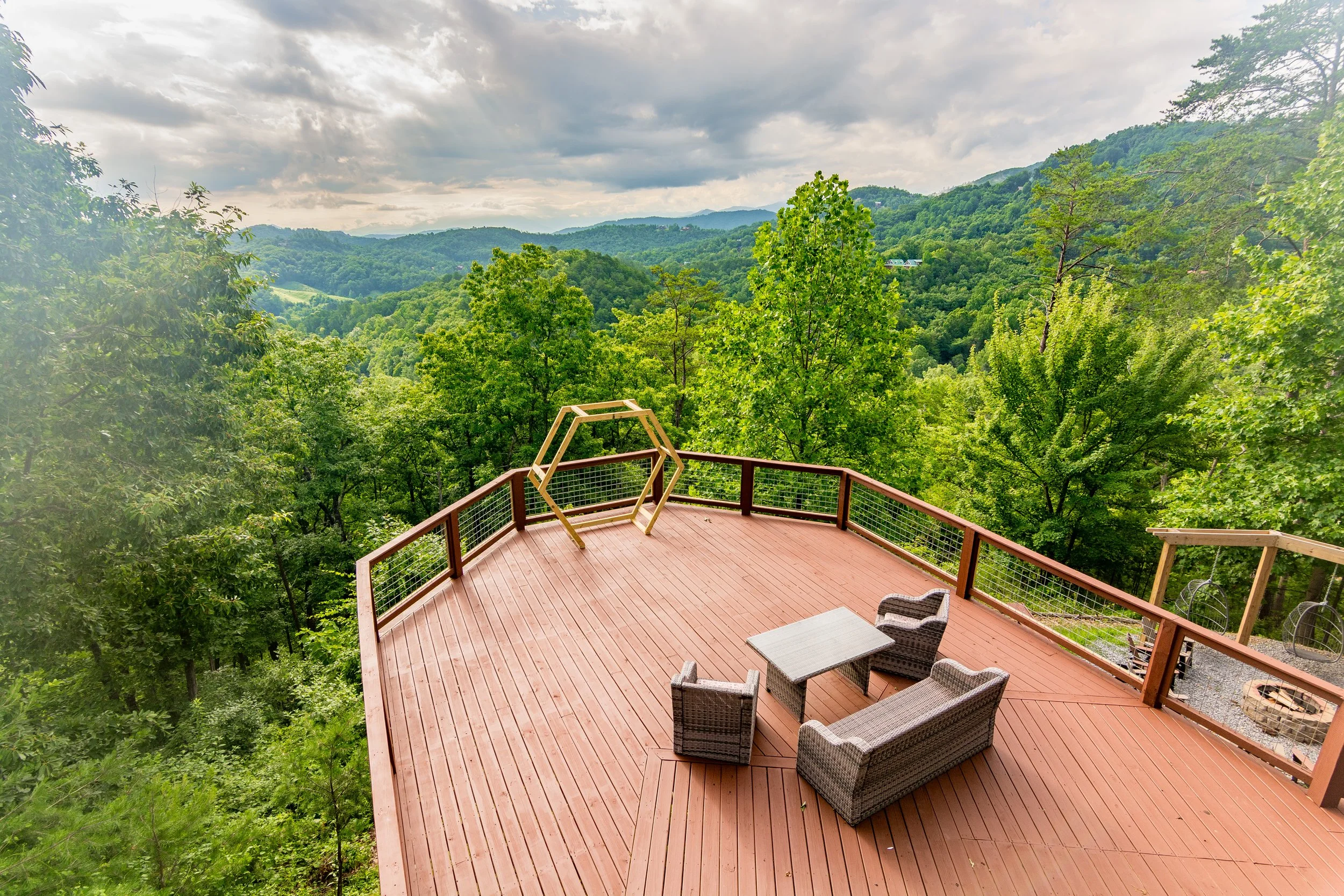 Wooden deck with outdoor furniture overlooking a lush green forested mountainscape under a cloudy sky.