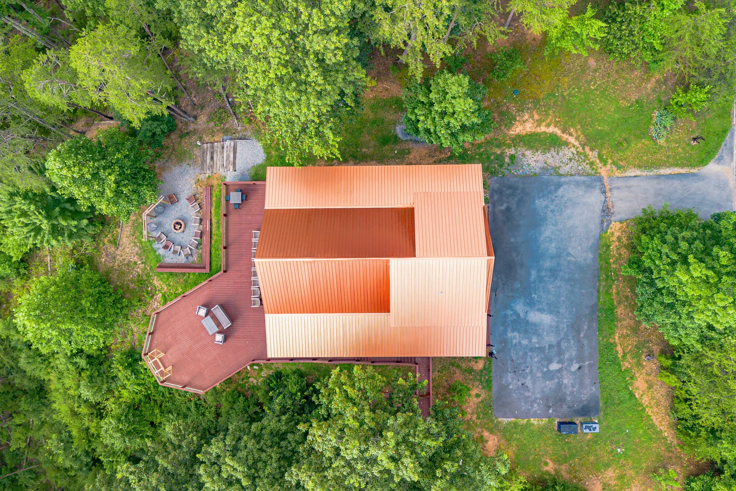 Aerial view of a house with a red roof, surrounding trees, a circular firepit with chairs, a deck, and a driveway.