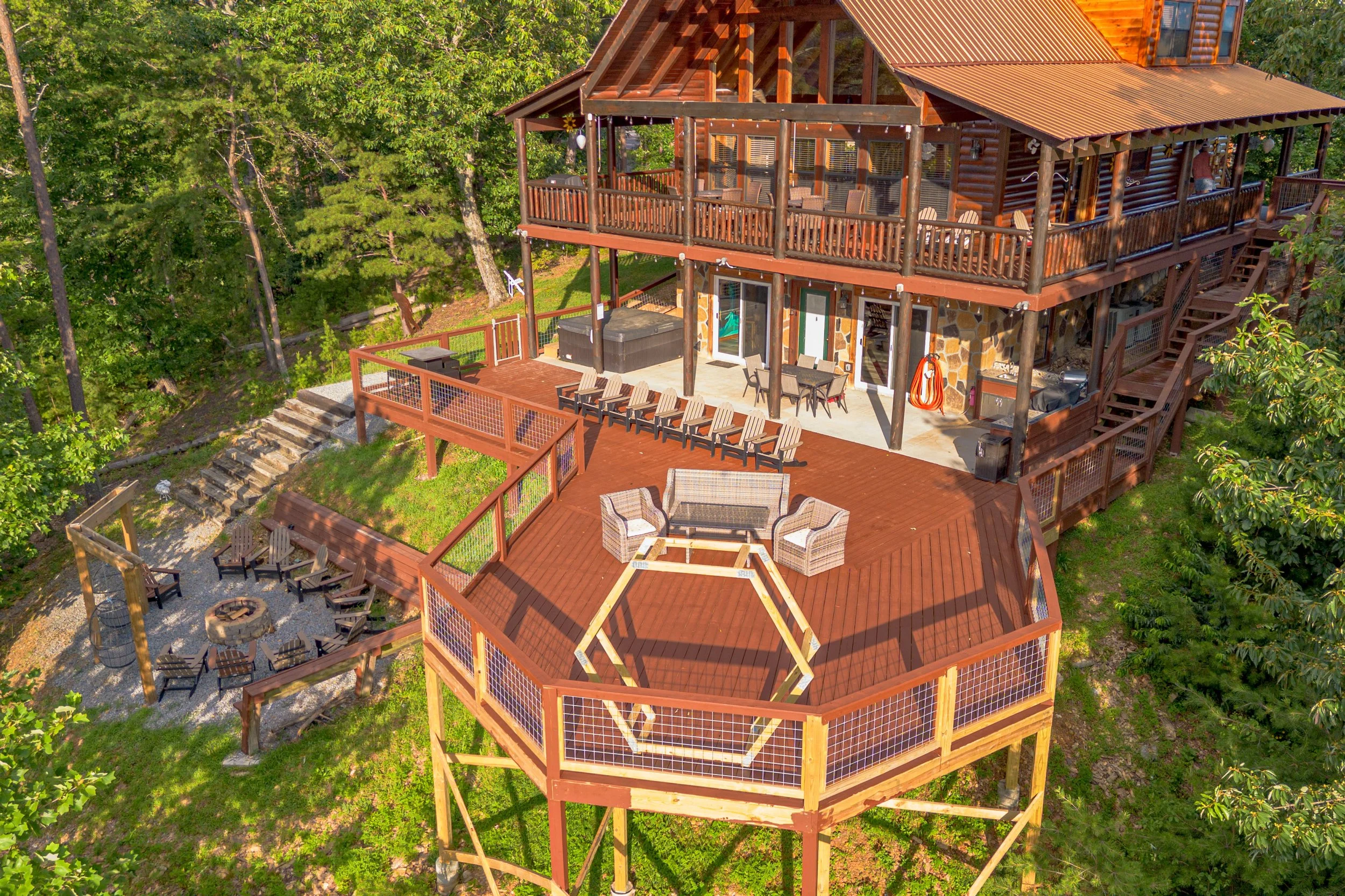 A top-down view of a multi-level wooden house with an expansive deck and a surrounding forest. The deck features multiple seating areas, including wicker chairs, a row of lounge chairs, a fire pit with chairs, and a hexagonal swing frame. Stairs lead