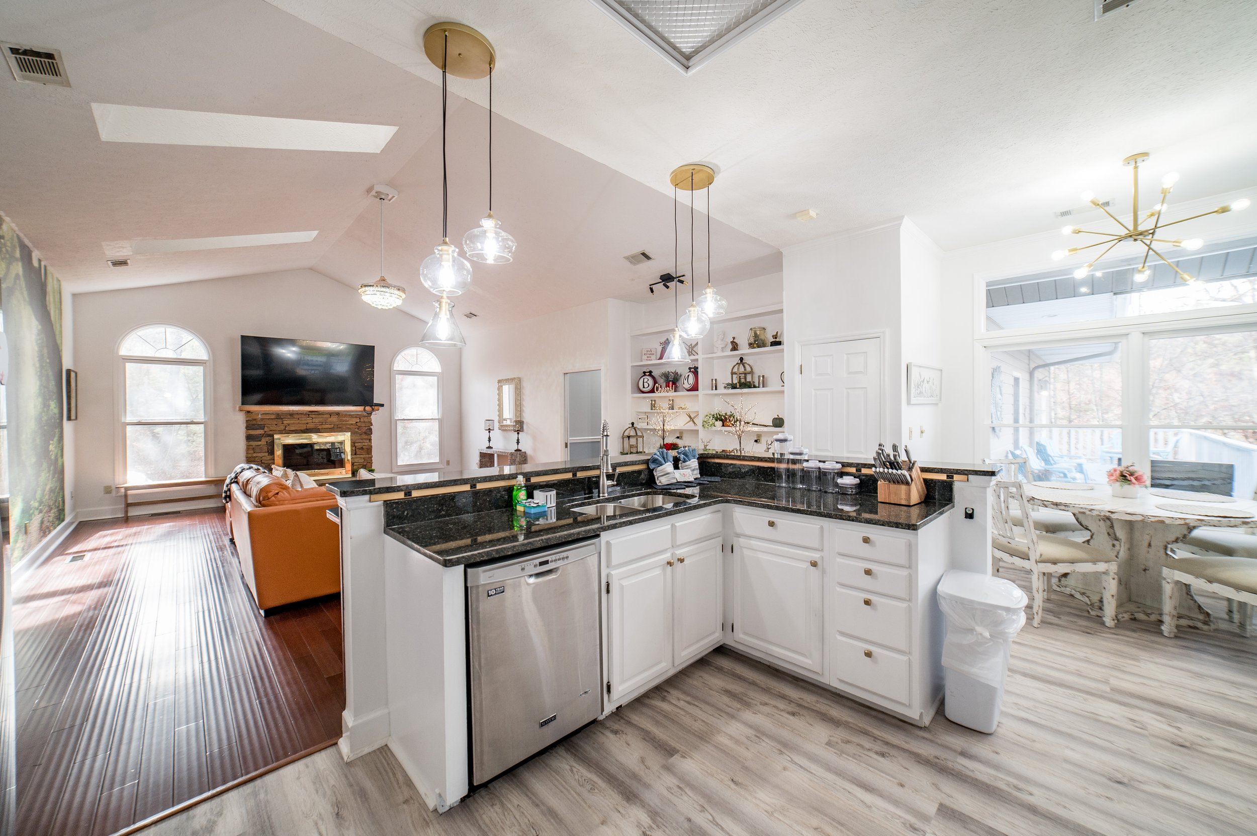 Open-concept living room and kitchen with white cabinets, black countertops, pendant lighting, a brick fireplace, a wall-mounted TV, and a dining area with large windows.