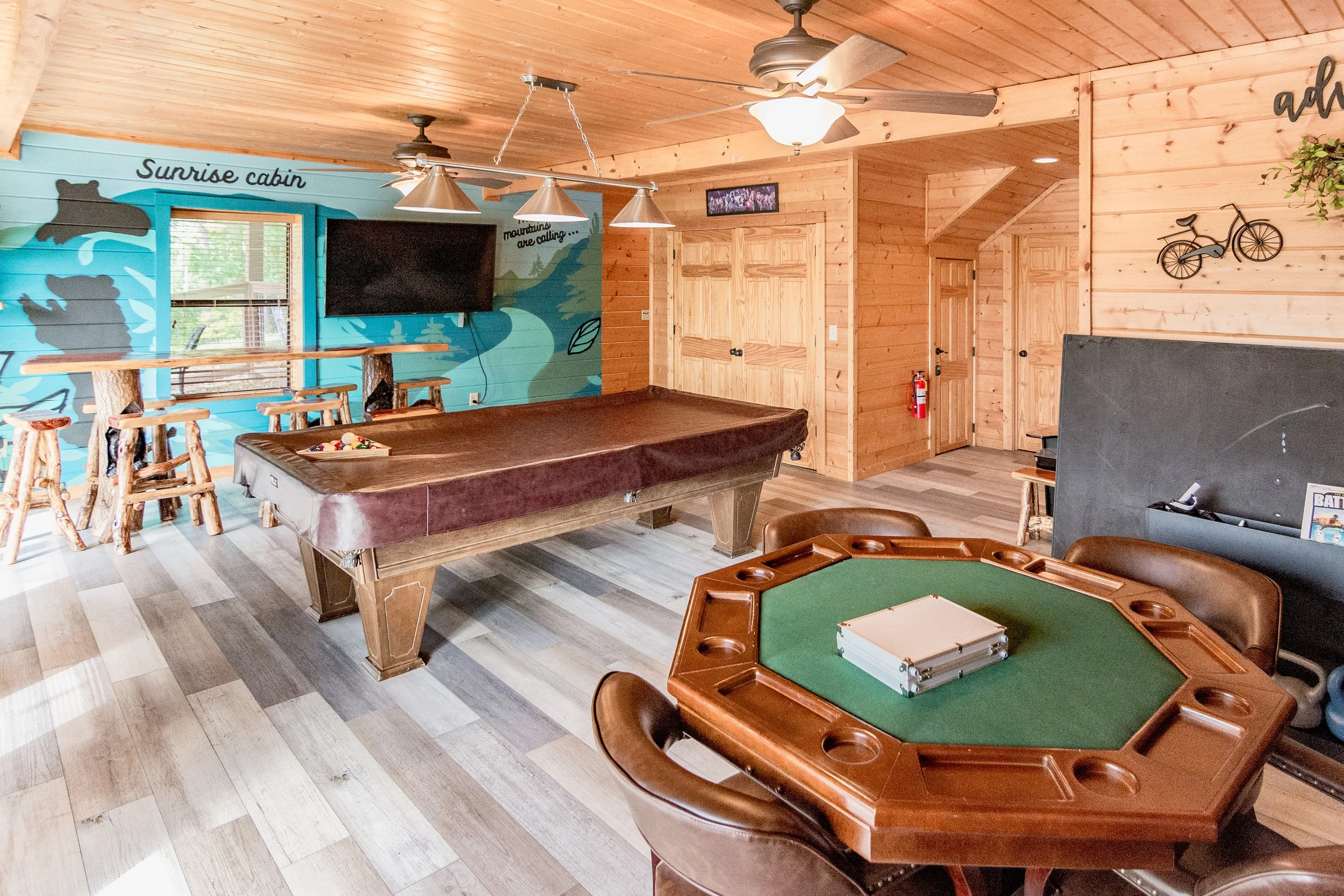 Interior of a wooden cabin game room with a pool table, a poker table, and a mural on the wall with the words "Sunrise cabin."