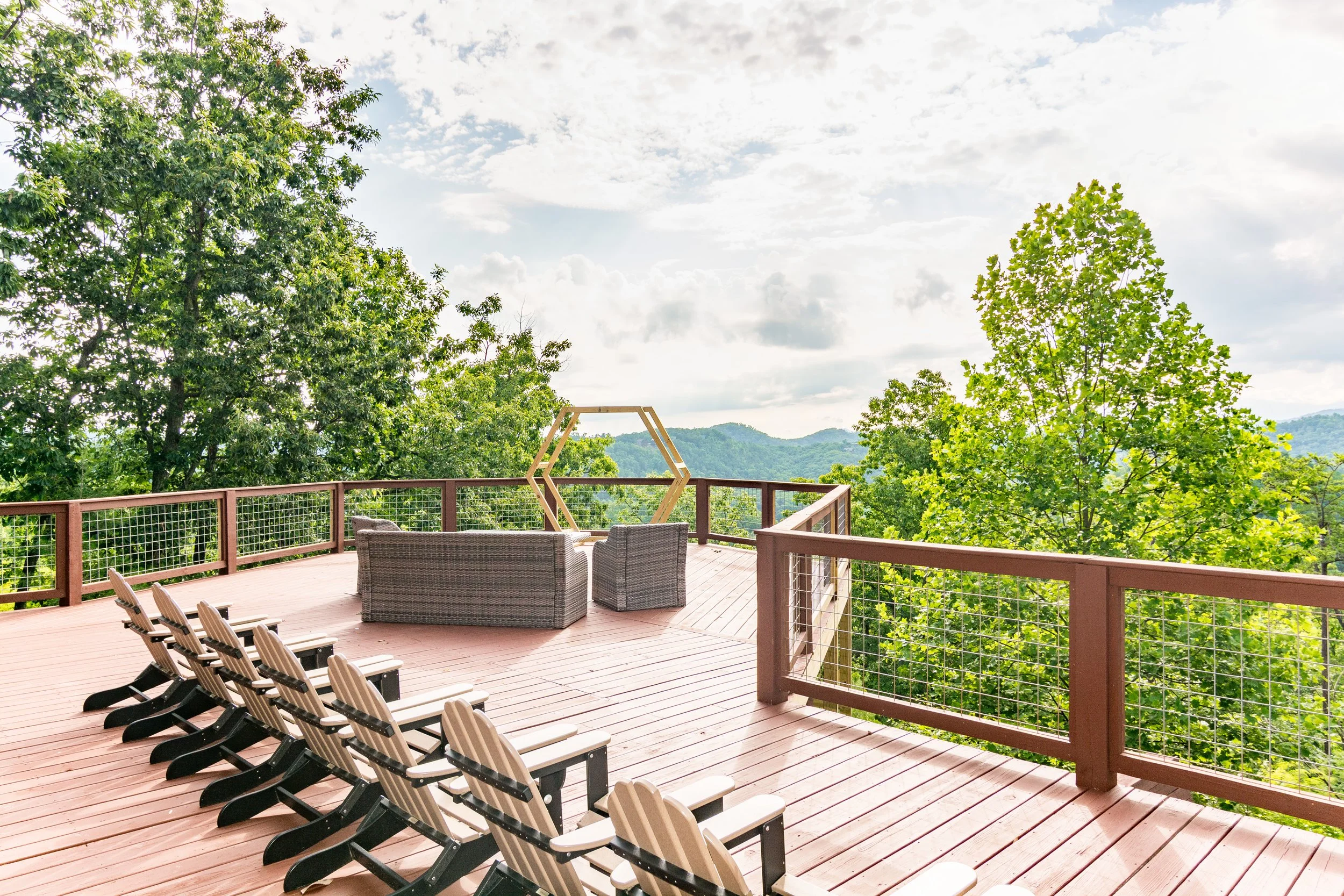 A spacious outdoor wooden deck with lounge chairs and outdoor furniture, overlooking a lush green forest with mountains in the background under a partly cloudy sky.