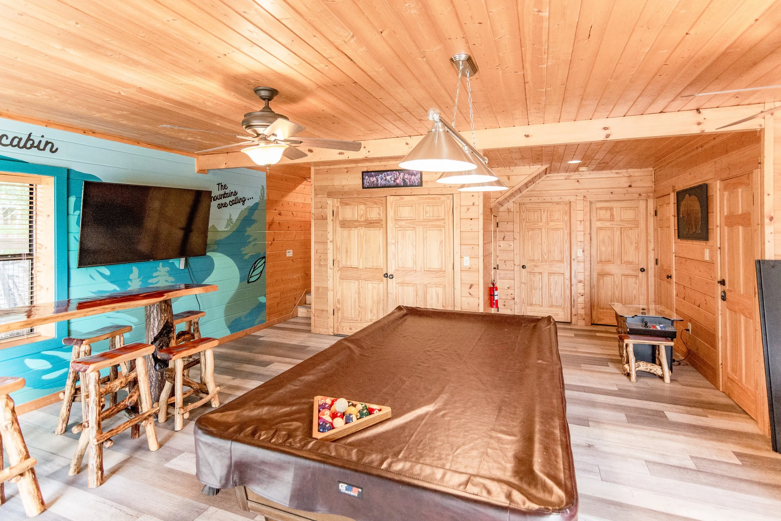 Interior of a cabin game room with a pool table, pool balls, and rustic wood decor, including a flat-screen TV on a colorful mountain mural wall.