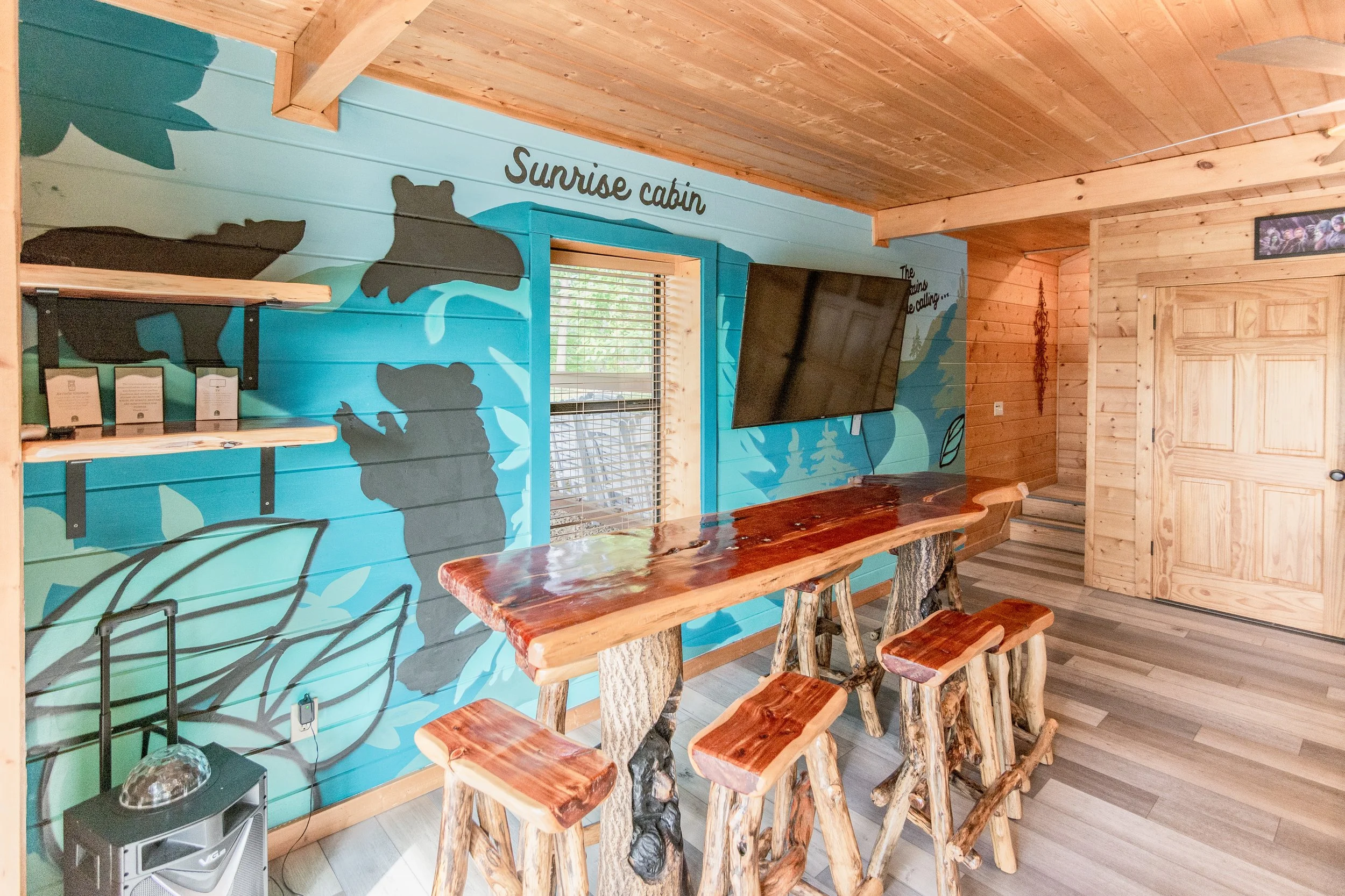 Interior of a rustic cabin with a painted wall featuring bears and the words "Sunrise cabin," a mounted flat-screen TV, a window with blinds, and a wooden table with stools made from tree branches.