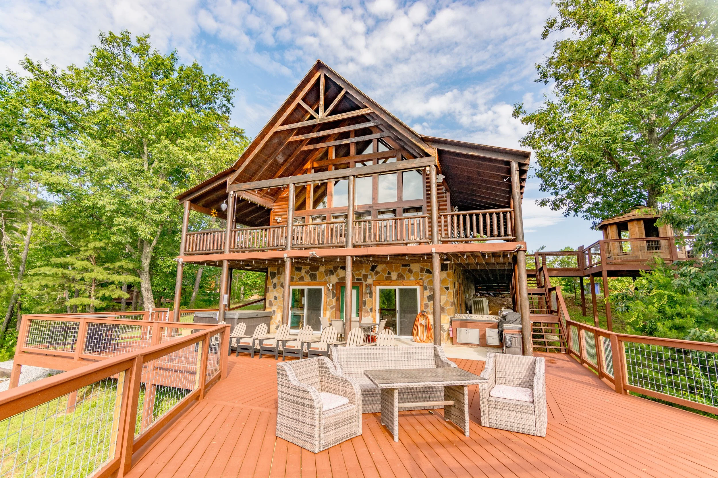 Large wooden cabin with multiple decks surrounded by green trees, outdoor furniture, and a stone wall on the lower level.