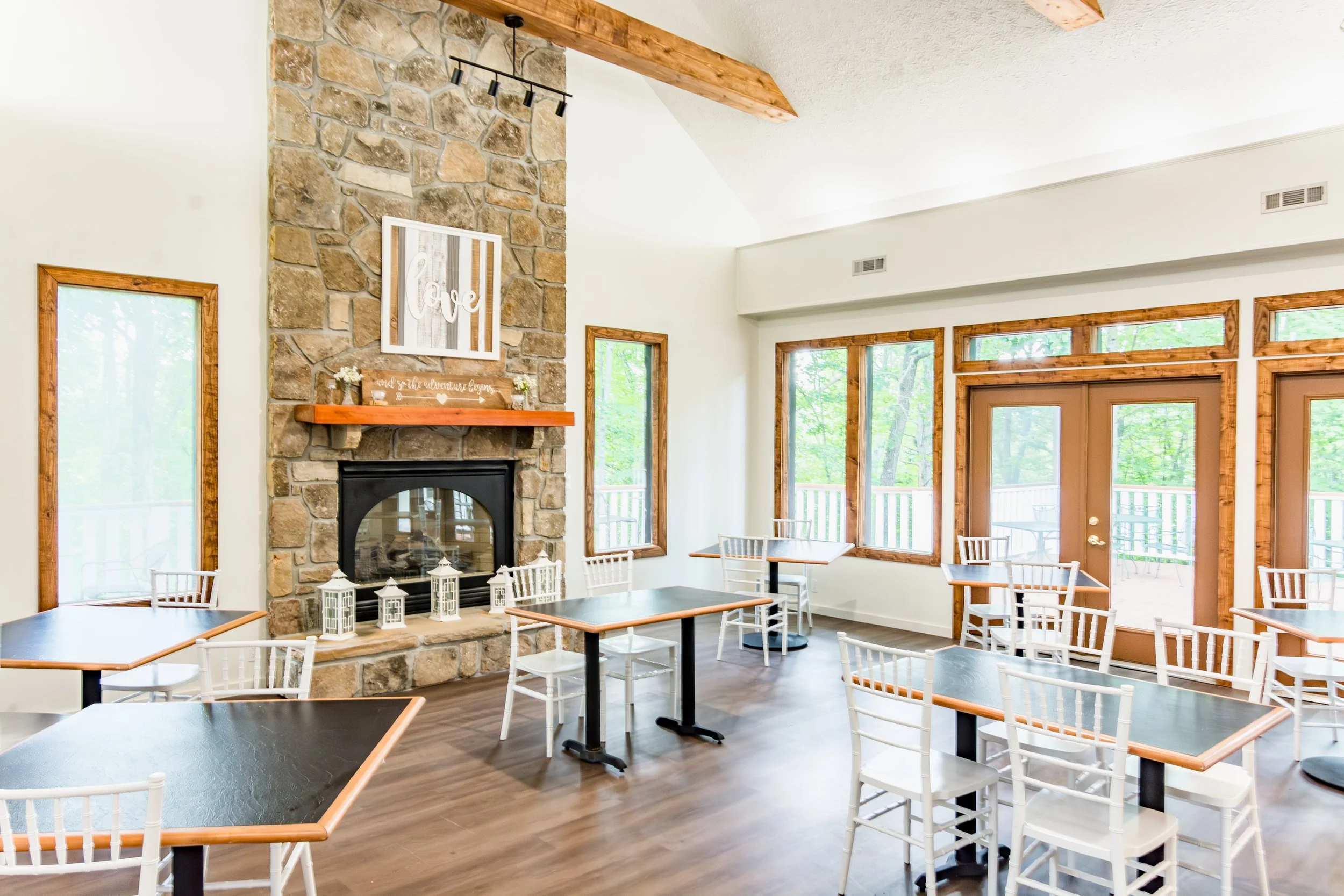 Bright dining area with tables and white chairs around a stone fireplace with decorative lanterns and love art above. Large windows and glass door reveal a wooded outdoor space.