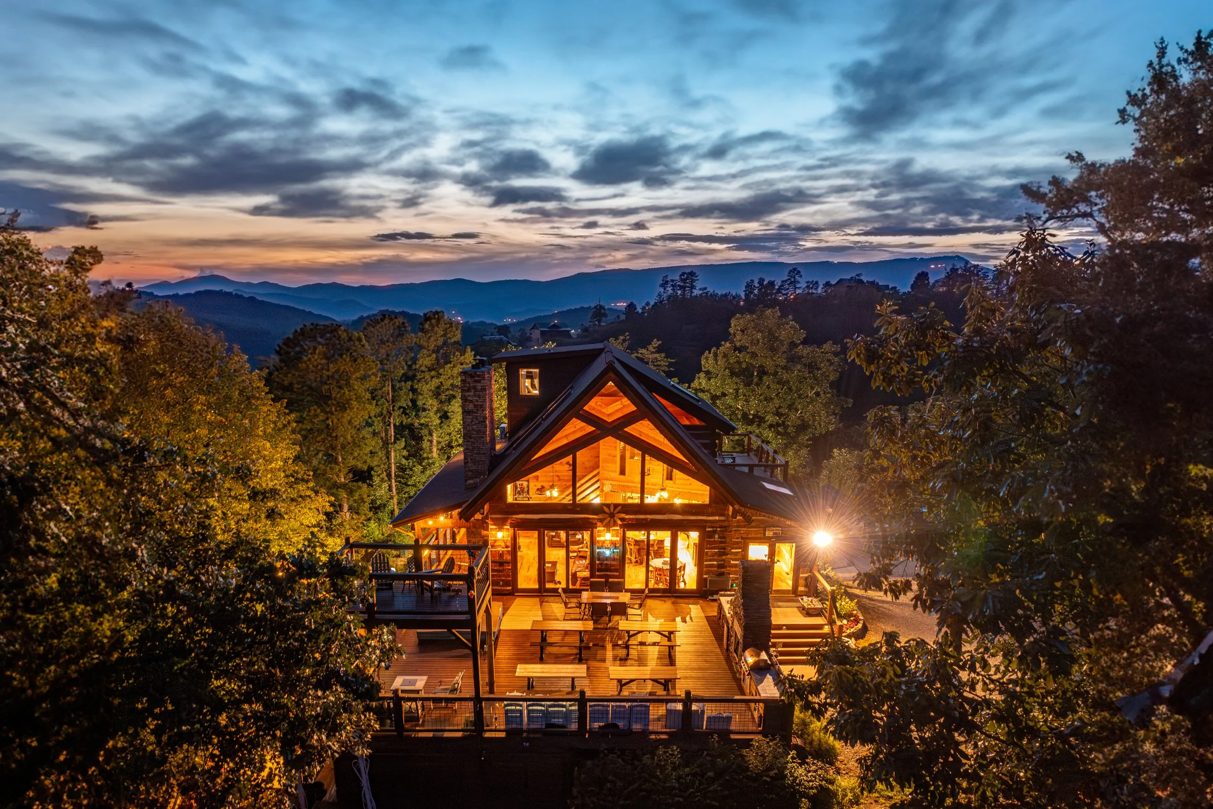 A cozy mountain cabin with large glass windows, illuminated from inside, surrounded by trees at dusk with mountains in the background.