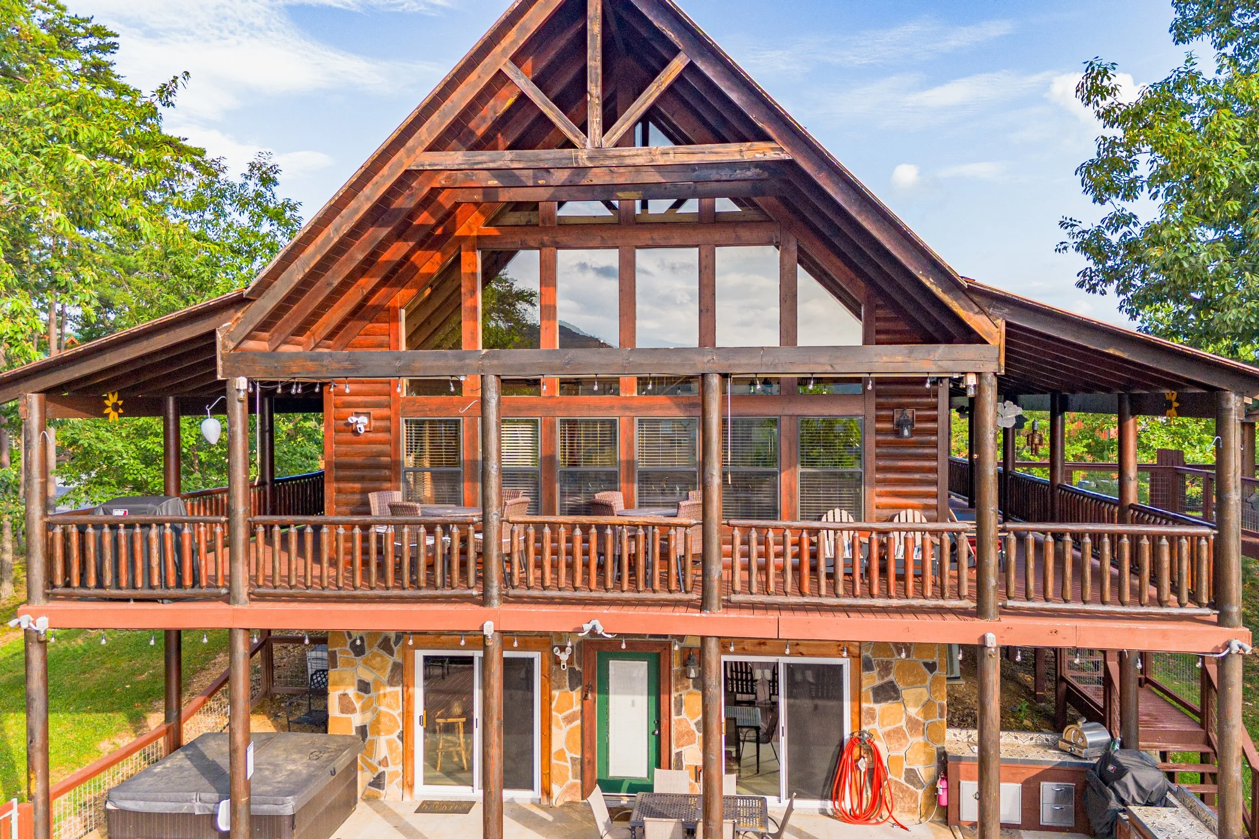 Large wooden house with two stories, a spacious wraparound deck on each level, wooden railings, and outdoor seating, surrounded by trees and a blue sky with clouds.