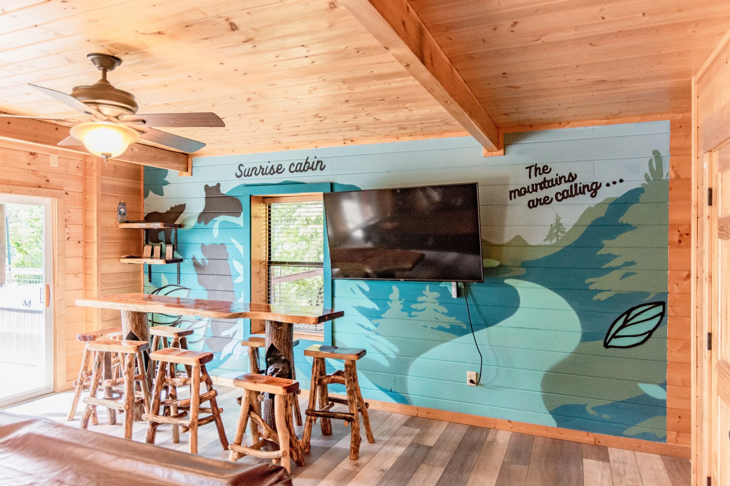 Interior of a wooden cabin with a wall mural depicting mountains and trees, a mounted flat-screen TV, a ceiling fan, and a seating area with a high wooden table and several stools.