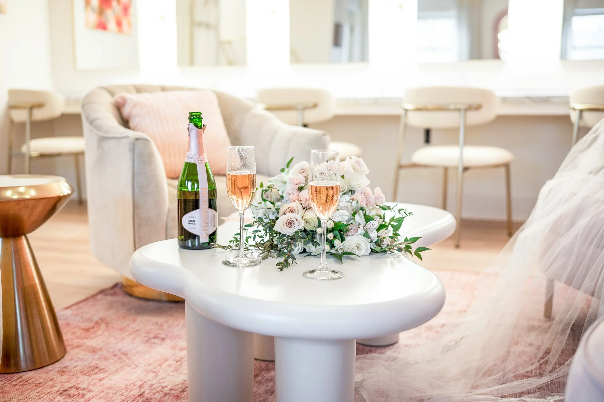 A white coffee table with a bouquet of flowers, a bottle of champagne, and two glasses of rosé wine in a bright living room with white furniture and pastel accents.
