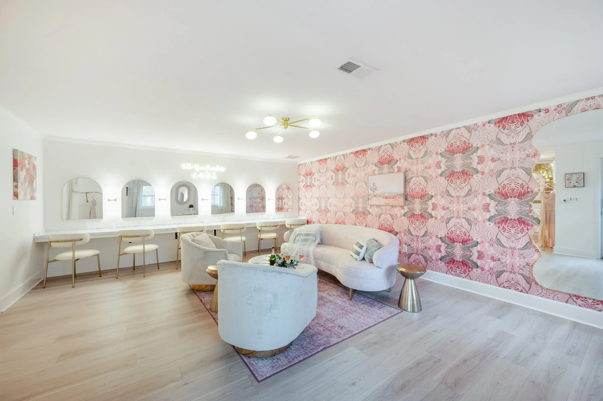 Bright living room with pink floral wallpaper, white curved sofa, two light blue chairs, a pink area rug, and a long white vanity with mirrors and chairs along the wall.