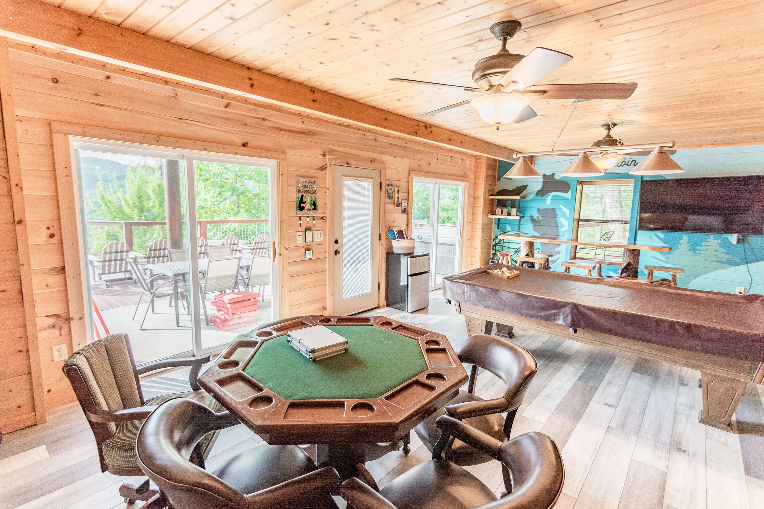 Interior of a wooden cabin room featuring a poker table, a pool table with cover, a mounted flat-screen TV, ceiling fans, and sliding glass door leading to an outdoor deck with chairs and tables.