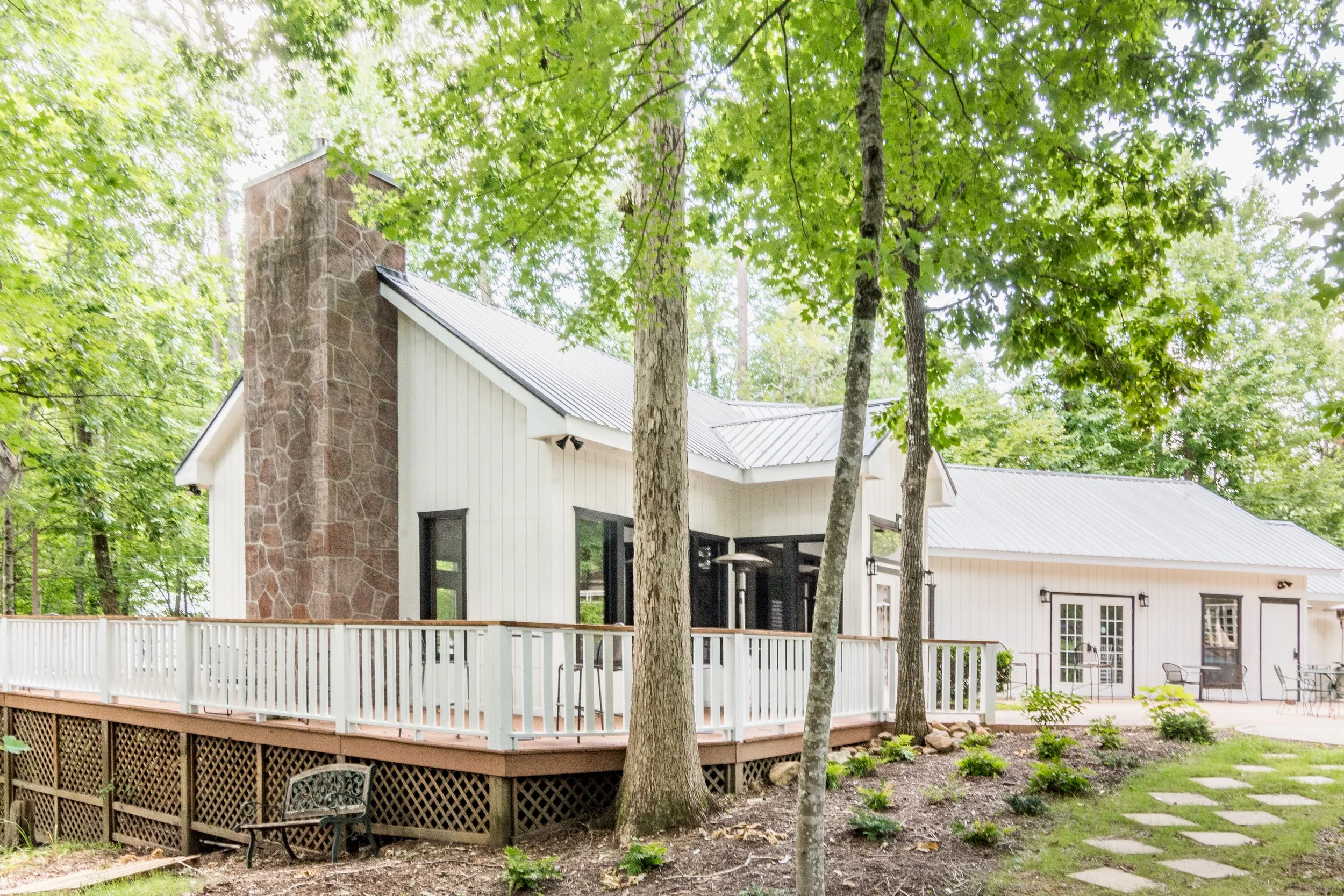 A modern house with white siding and a stone chimney, surrounded by trees and a yard with stone pathway and outdoor patio furniture.