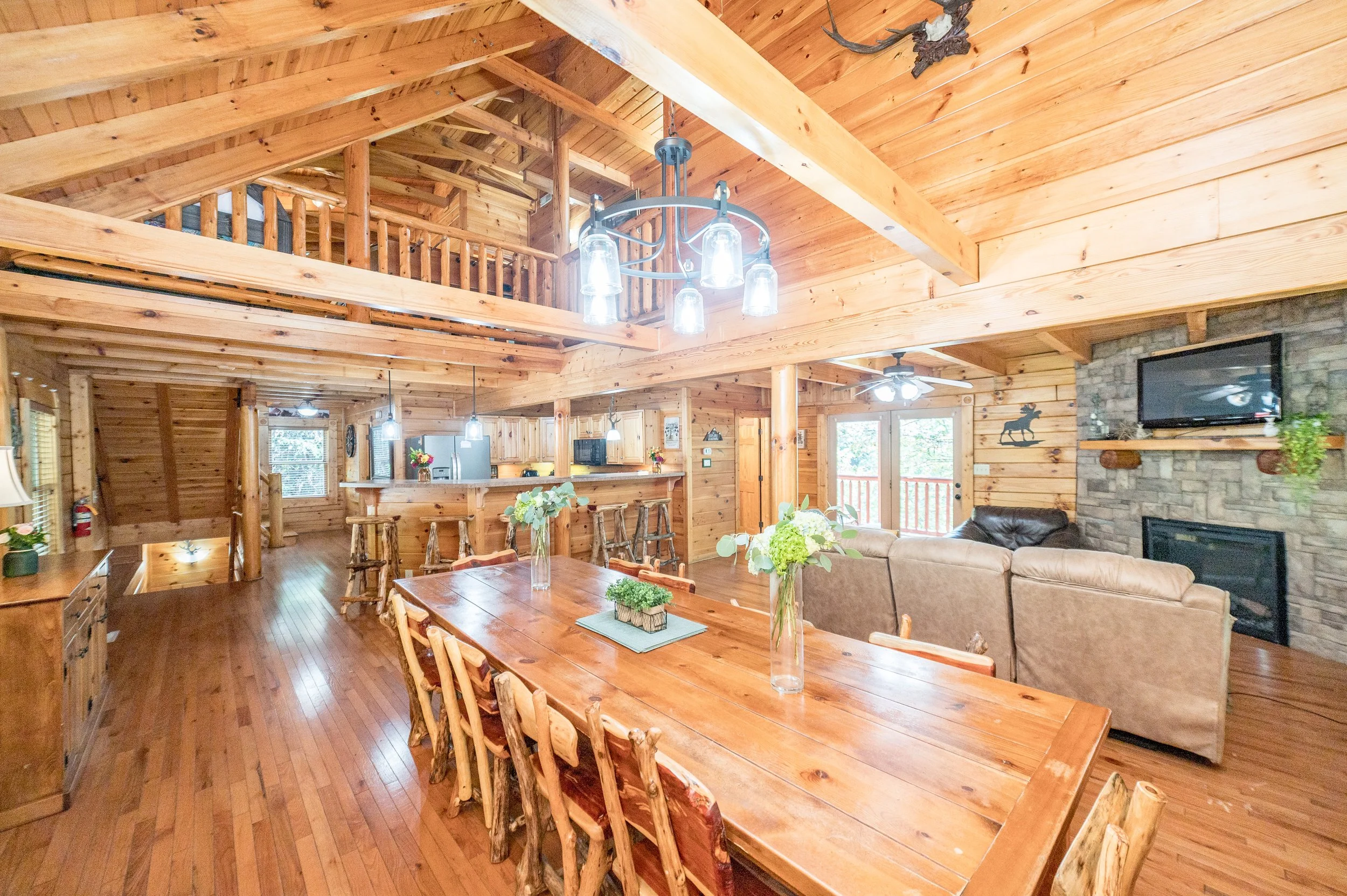 Interior of a cozy wooden cabin living room and dining area with a large wooden dining table, floral centerpieces, a stone fireplace with a mounted TV, and a kitchen in the background, featuring wood paneling and rustic decor.