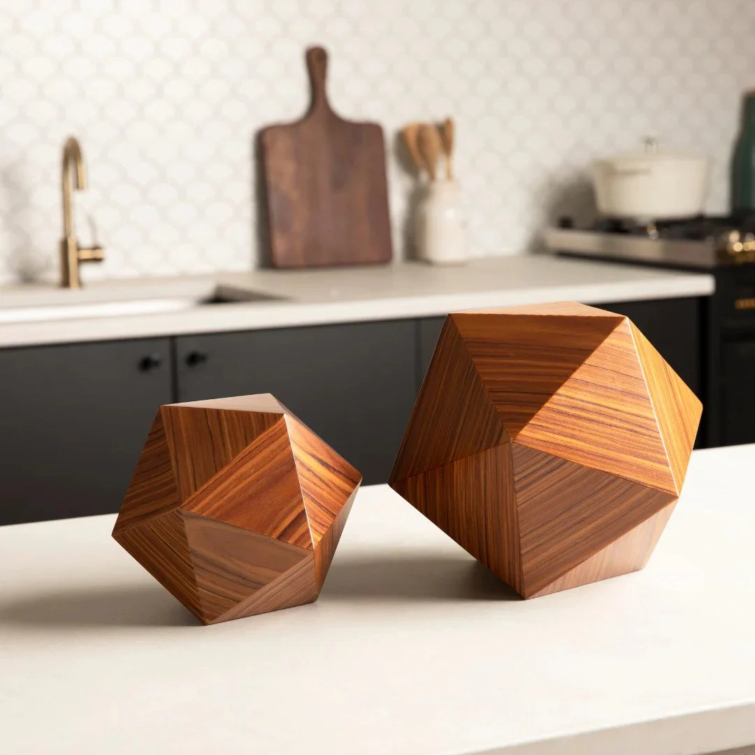 Two wooden geometric sculptures on a white countertop in a modern kitchen with dark cabinets, a white backsplash, and kitchen utensils in the background.