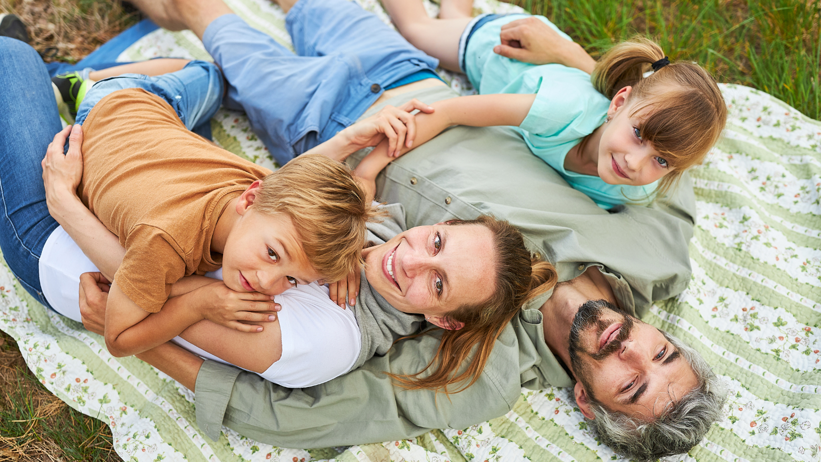 Une famille (papa, maman, garçon et fille) profitent du soleil allongés sur une couverture dans l'herbe