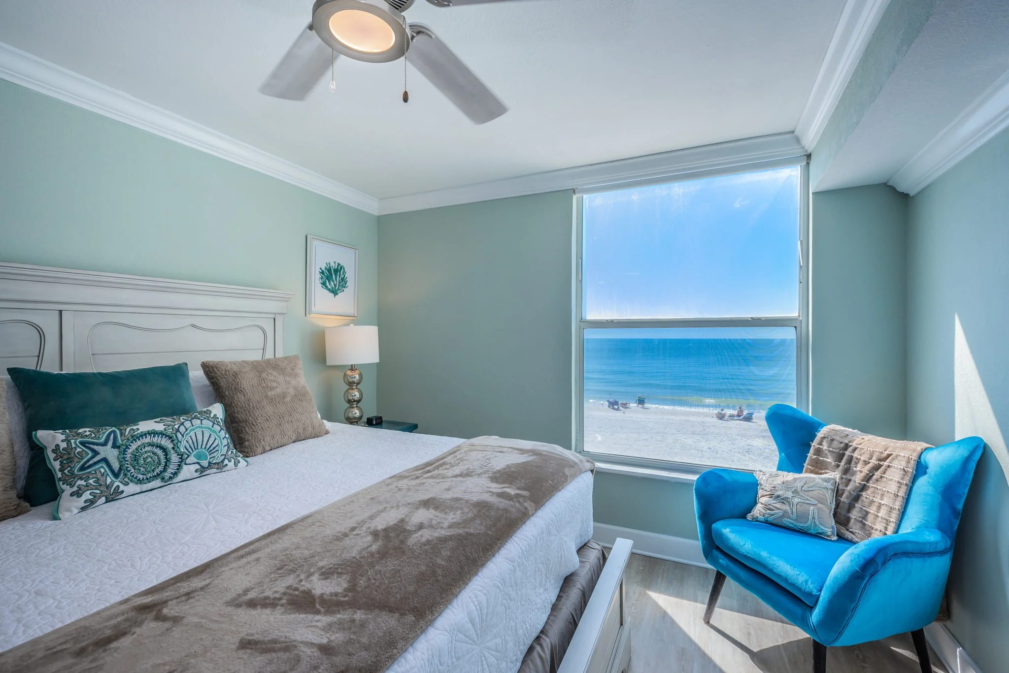 Beachside bedroom with a large window showing the ocean, a white bed with patterned pillows, a bedside lamp, and a bright blue armchair near the window.