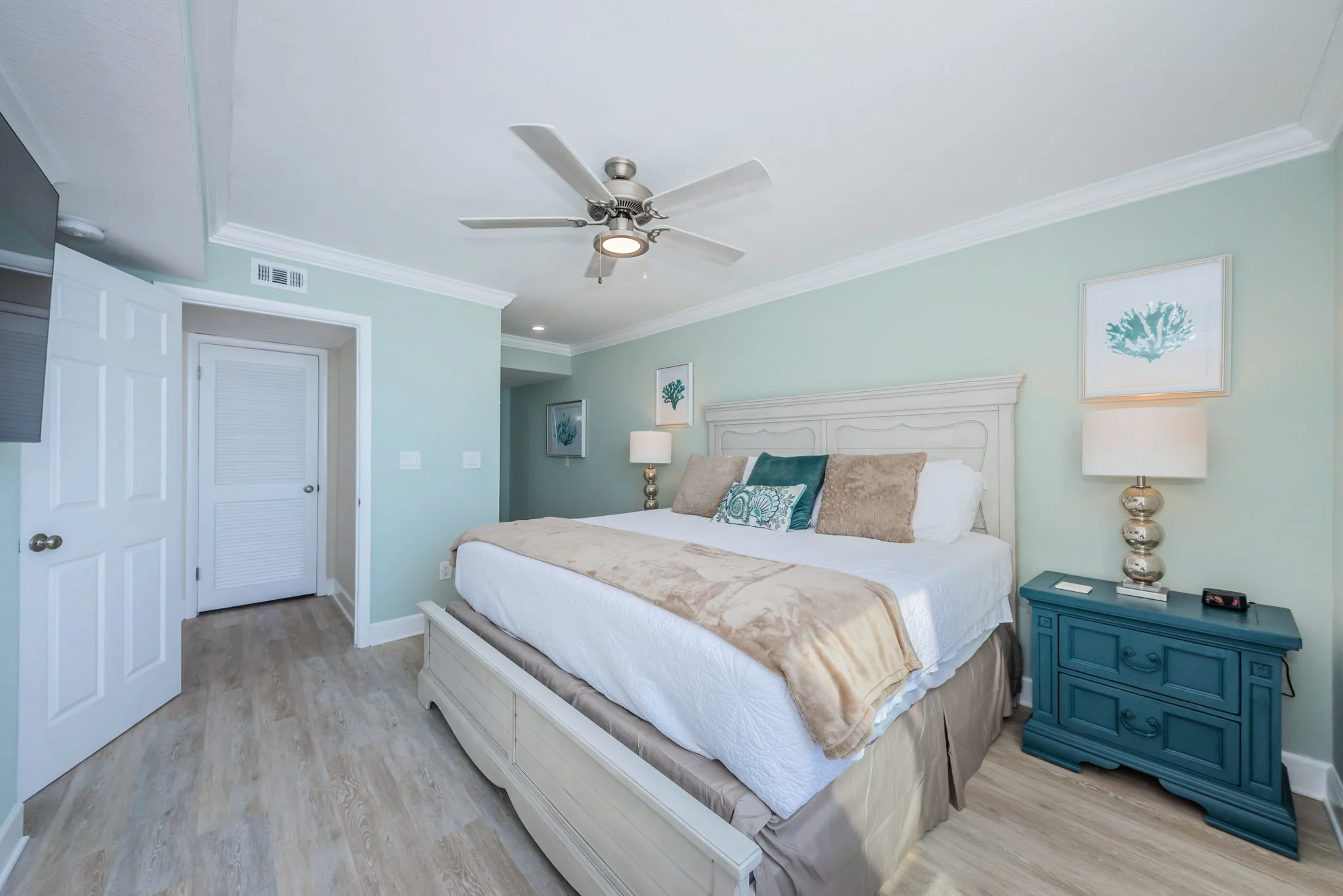 A coastal-style bedroom with a king-sized bed, light green walls, white crown molding, a ceiling fan, two bedside tables with lamps, and framed artwork of coral.