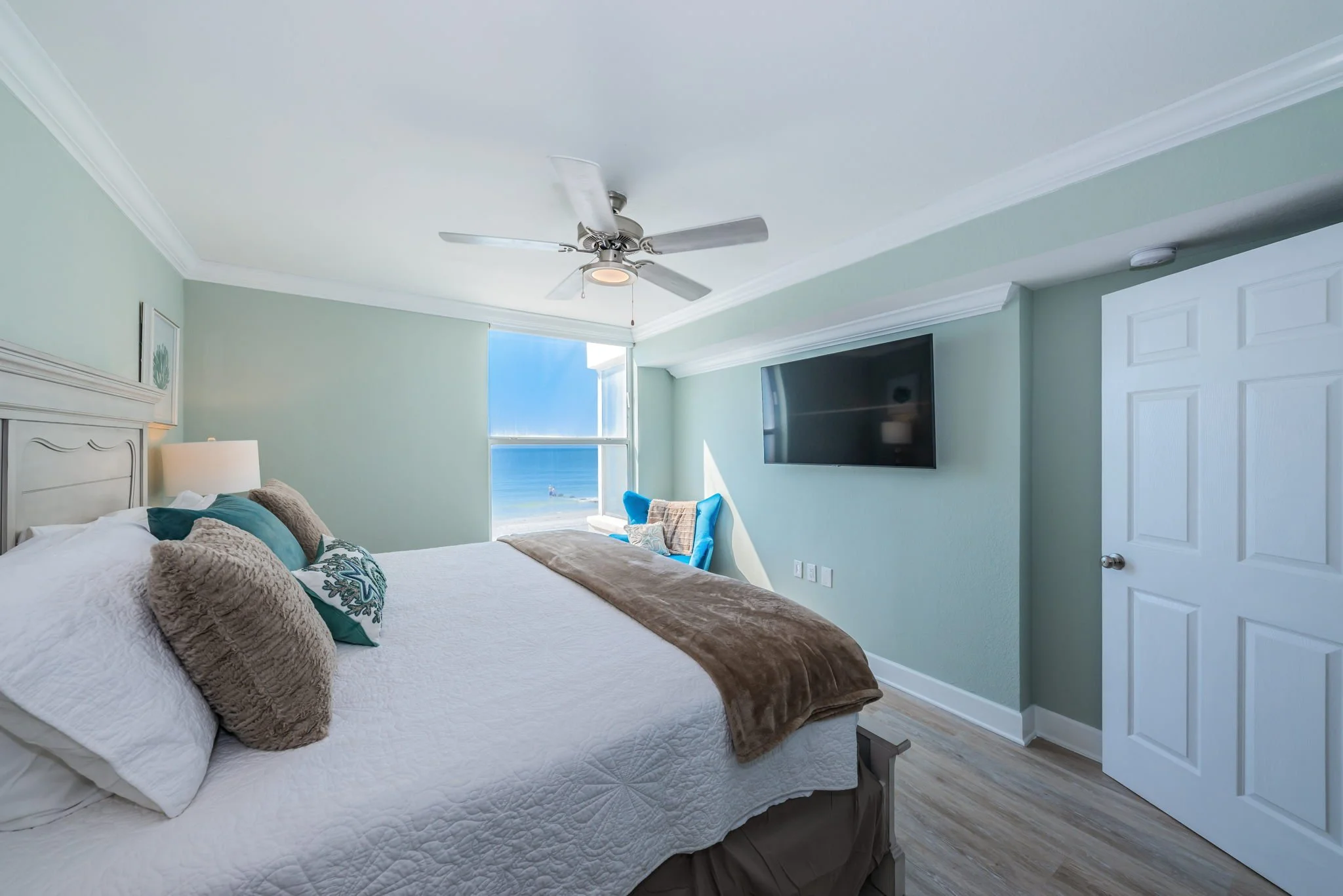 Bedroom with large bed, ocean view window, wall-mounted TV, ceiling fan, light green walls, door, and light wood flooring.