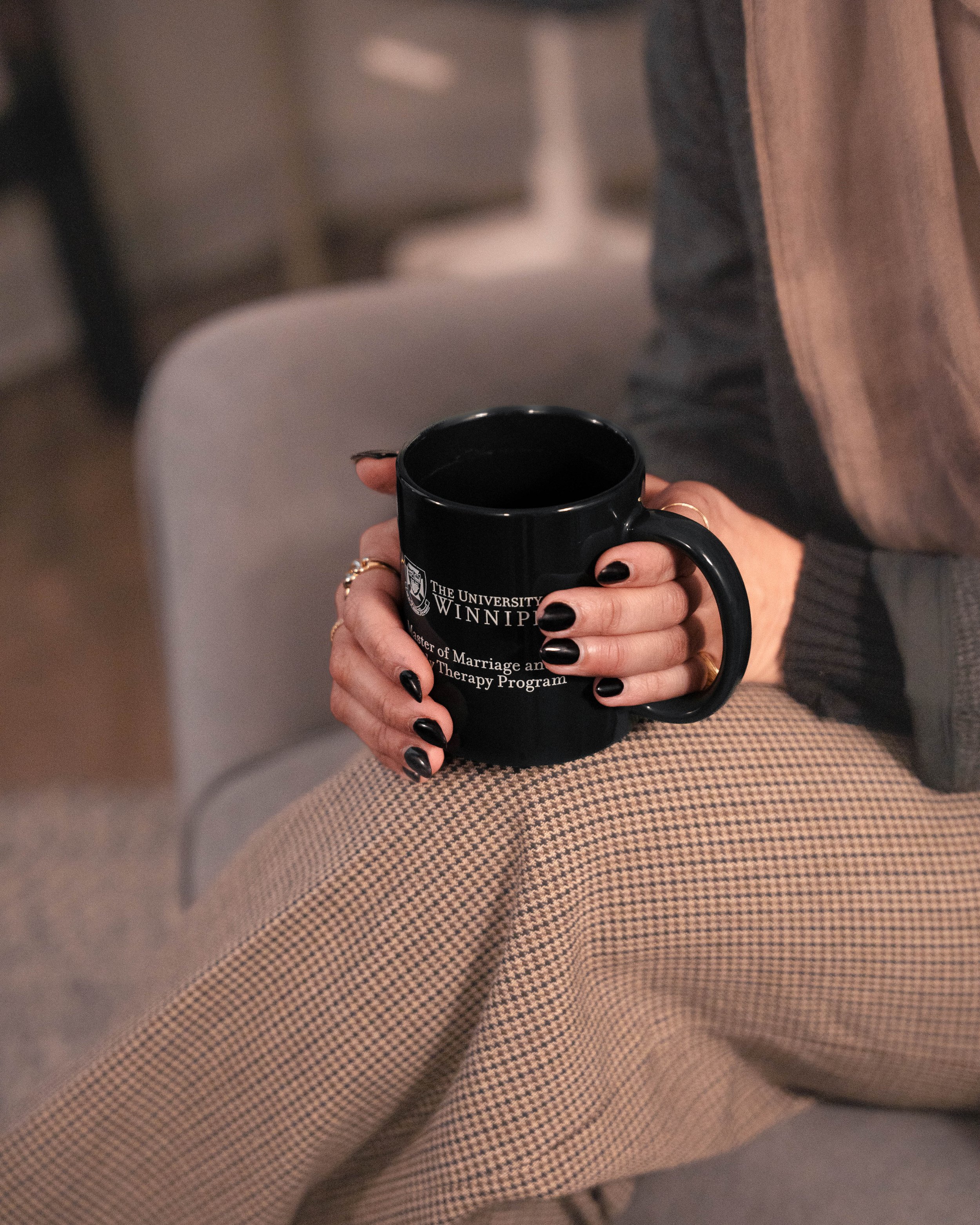 Person holding a black ceramic mug with University of Winnipeg logo and text on it, sitting on a beige and brown checkered fabric surface, wearing beige checked pants and a gray jacket.