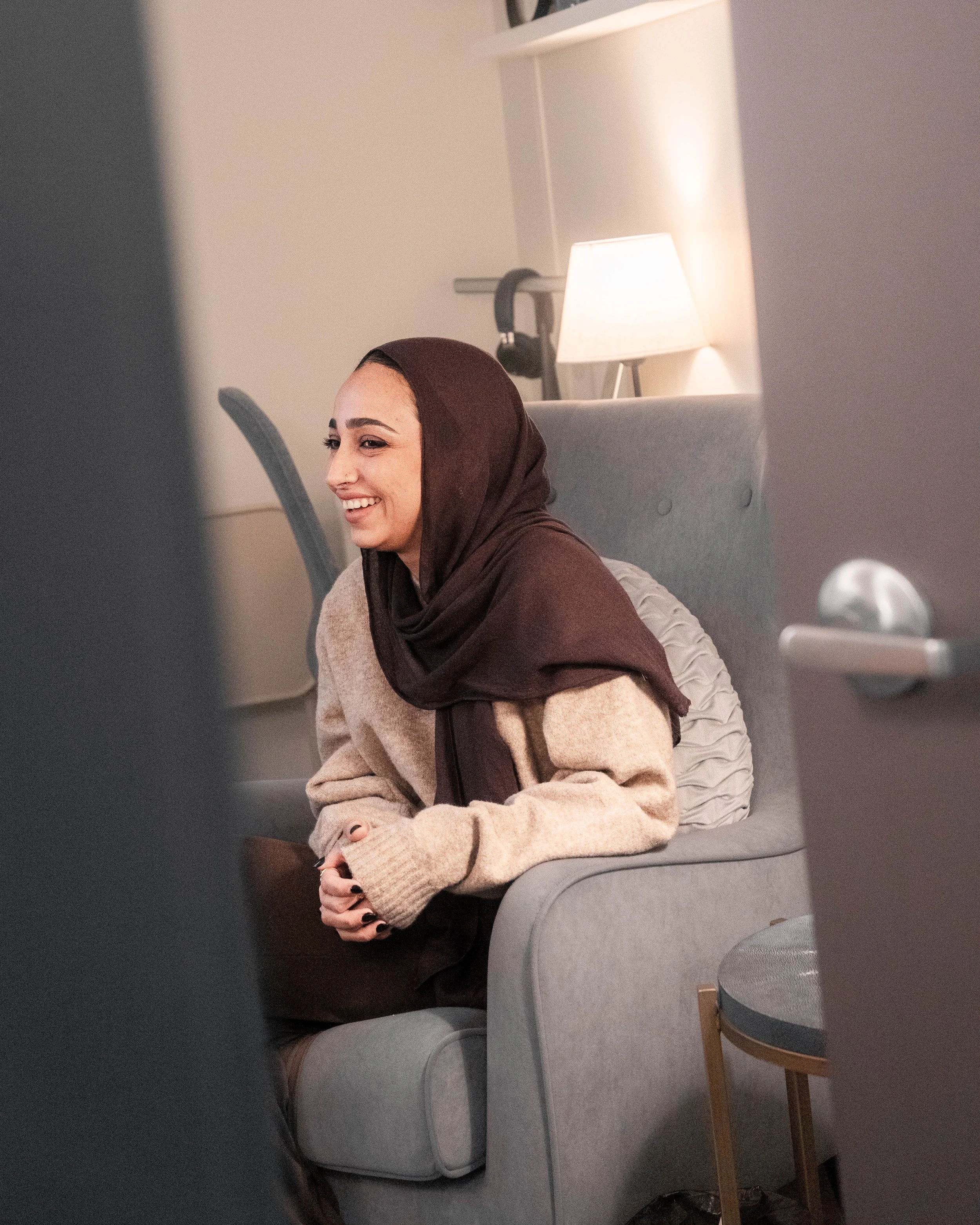 A woman sitting on a gray armchair, smiling, wearing a brown hijab and a beige sweater, in a cozy room with a lamp and a white pillow behind her.