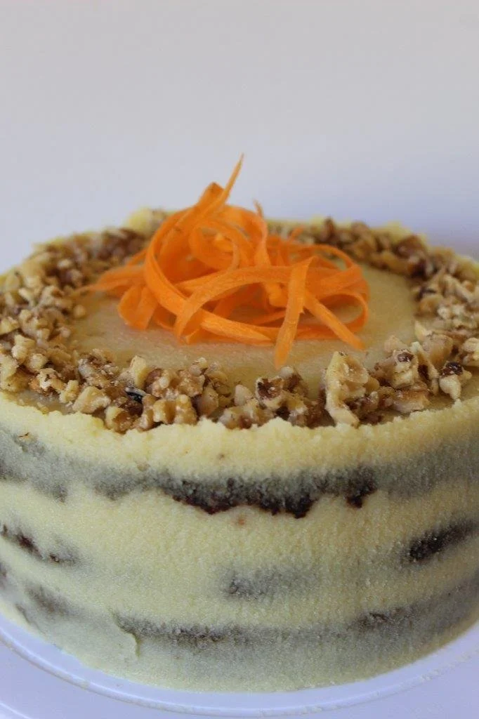 Close-up of a layered cake with cream-colored frosting, topped with shredded carrots and chopped nuts around the edge.