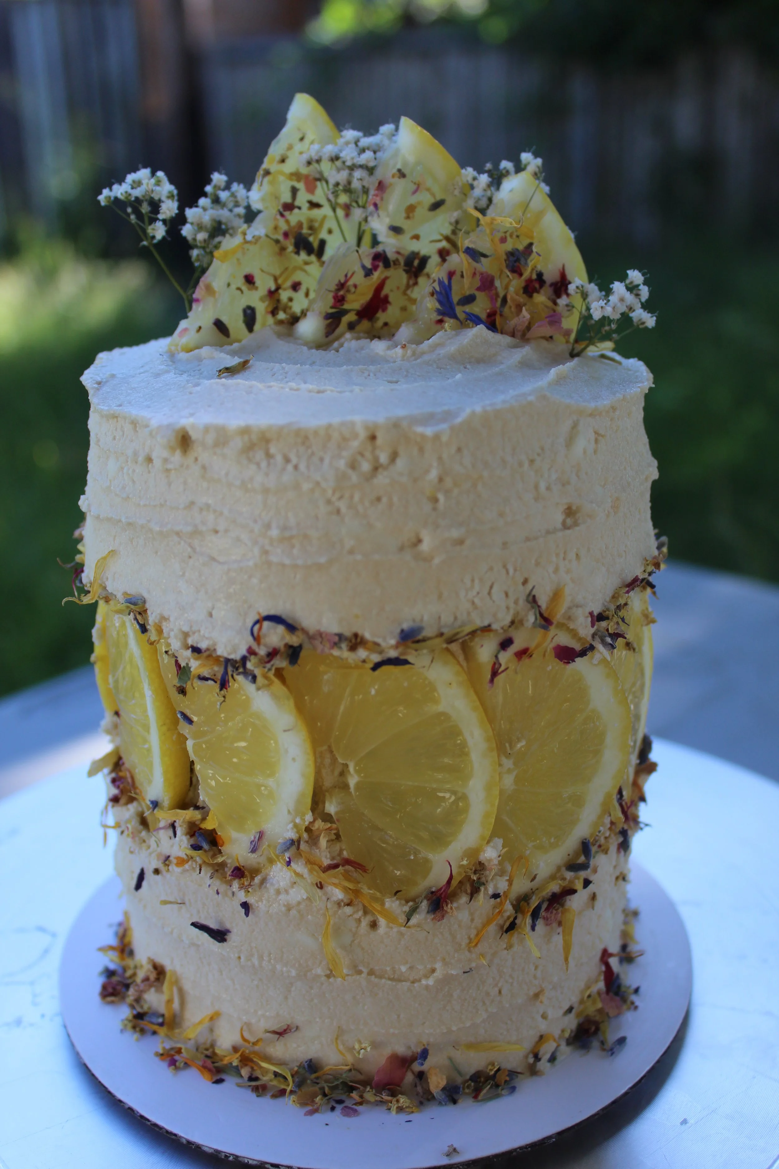 A two-tiered lemon-flavored cake decorated with lemon slices, edible flowers, and white frosting, set outdoors.