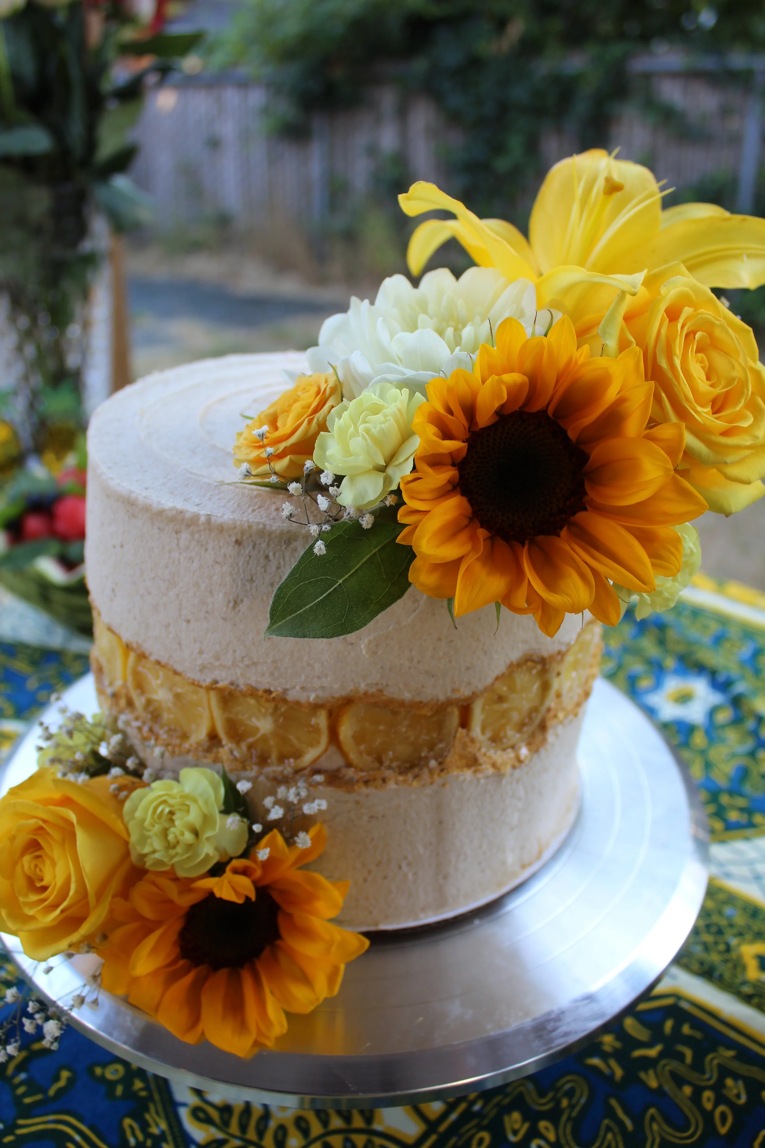 A two-tiered cake decorated with fresh yellow roses, sunflowers, white flowers, and greenery, set on a silver cake stand outdoors with a blue and yellow patterned tablecloth.