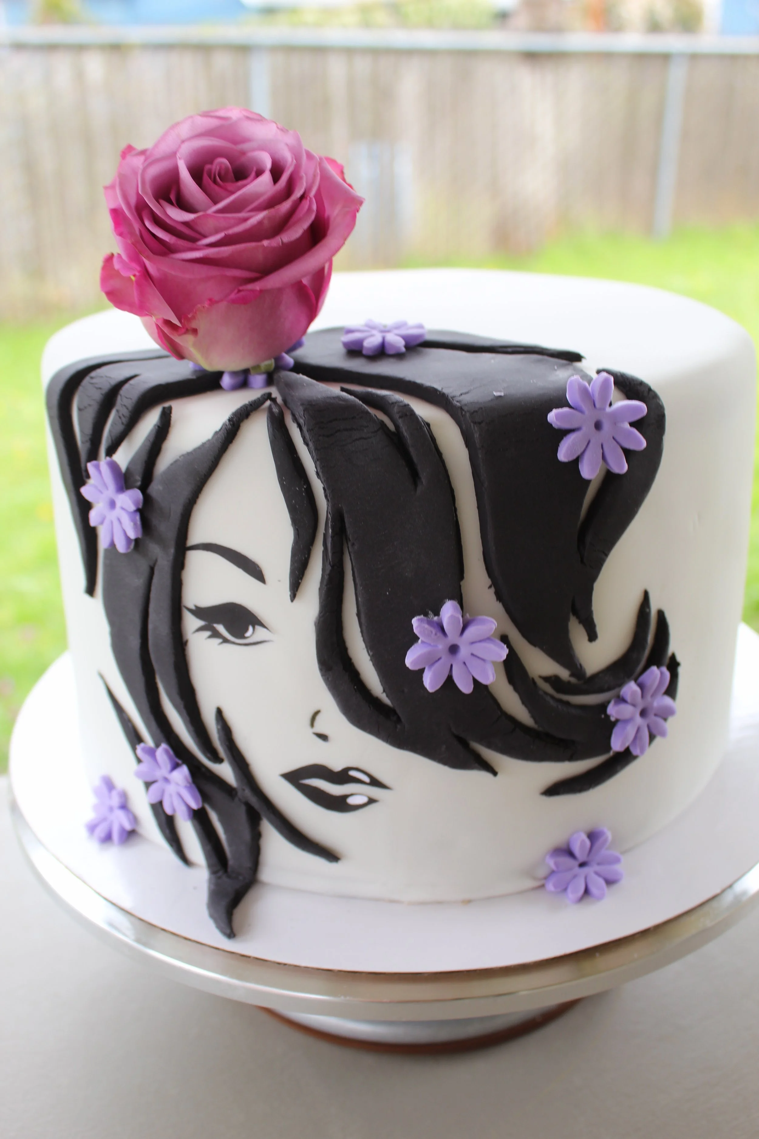 A round white cake decorated with black and white icing in the shape of a woman's face with black hair and purple flowers, topped with a pink rose.