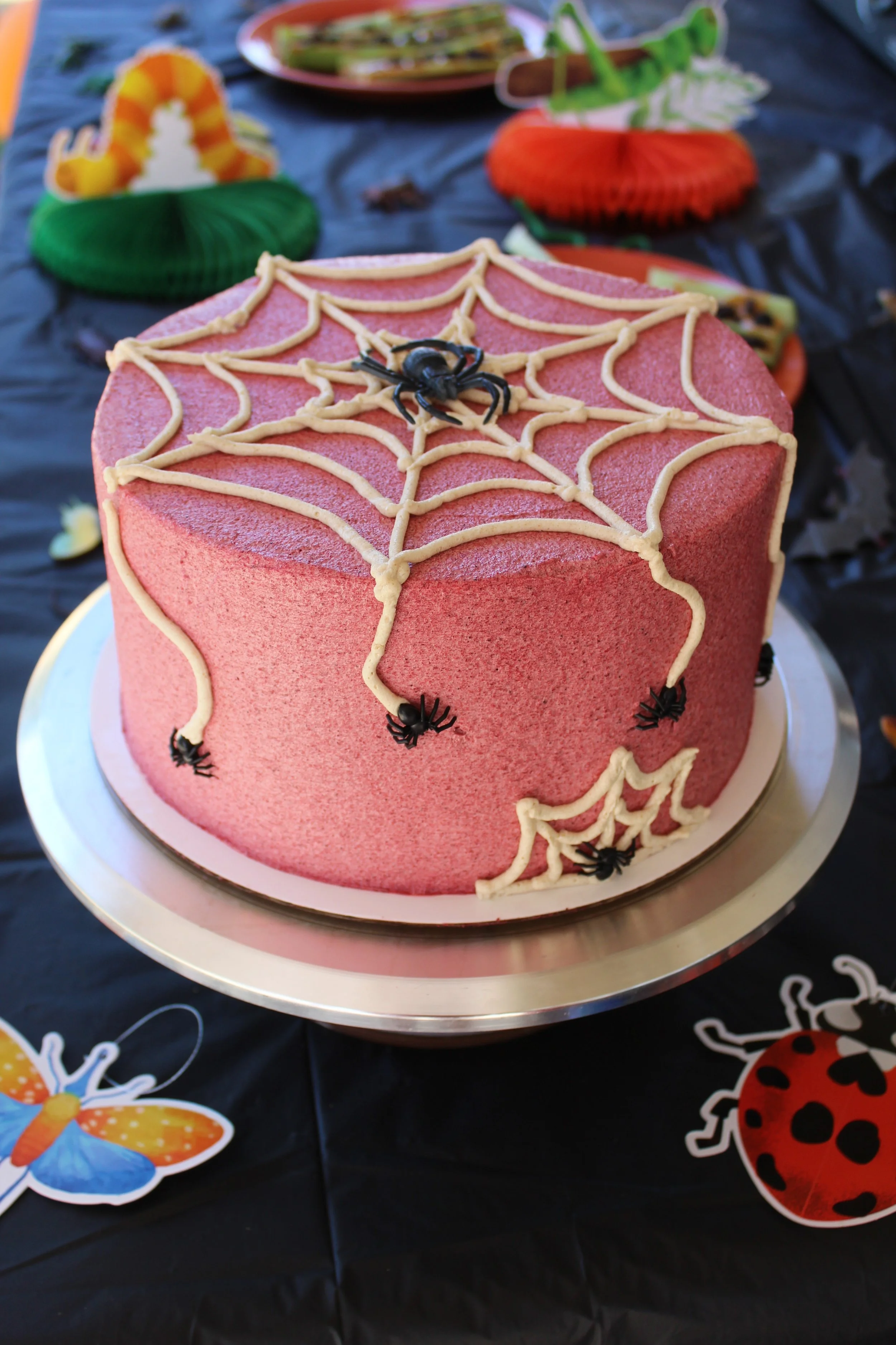 A pink Halloween-themed cake decorated with a spider web design made of icing and small plastic spiders, placed on a silver cake board. The cake is on a black table with seasonal decorations, including a butterfly and a ladybug cutout.
