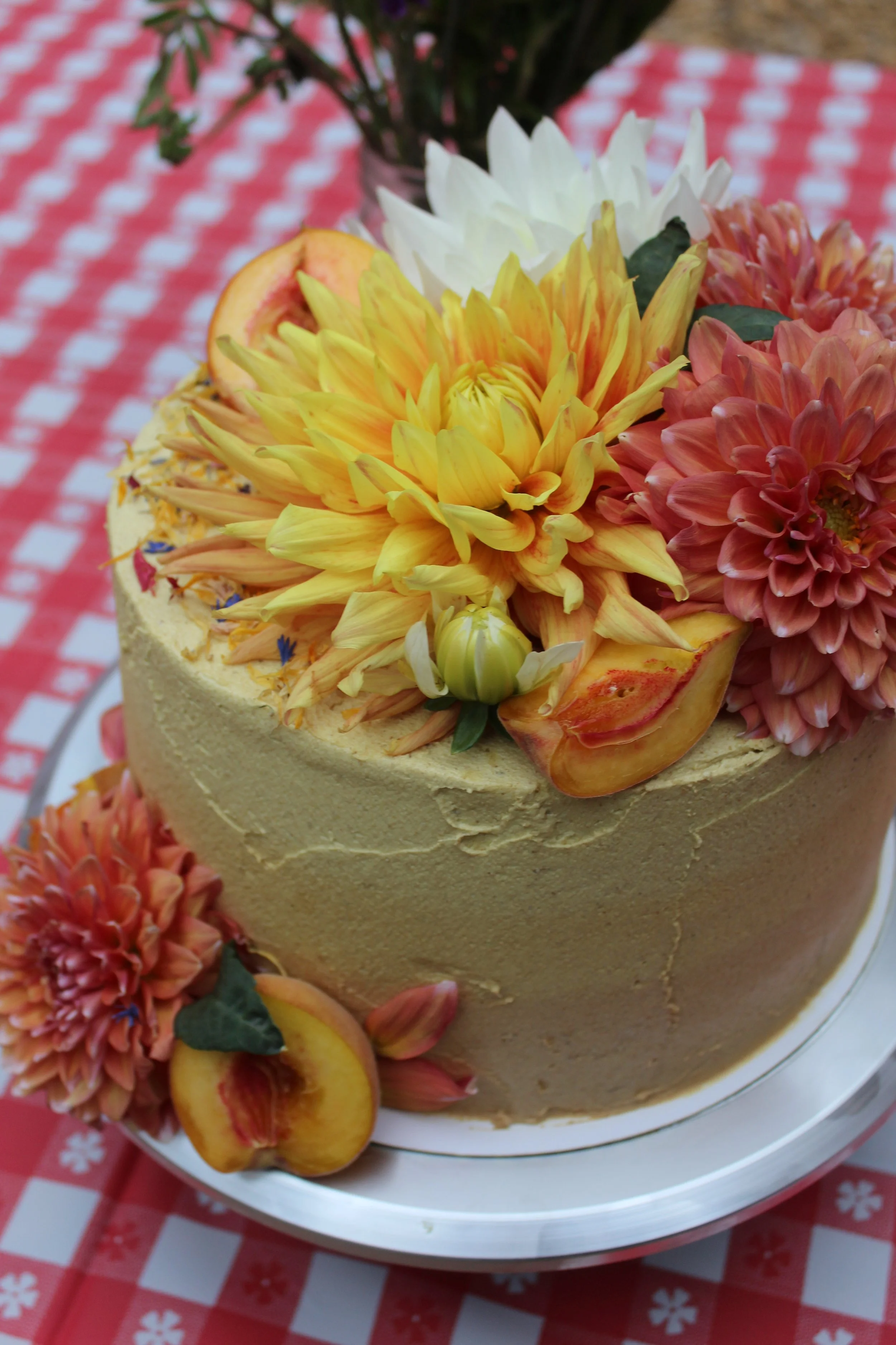 Decorated cake with yellow, pink, and white flowers, peaches, and flower petals on a red checkered tablecloth.